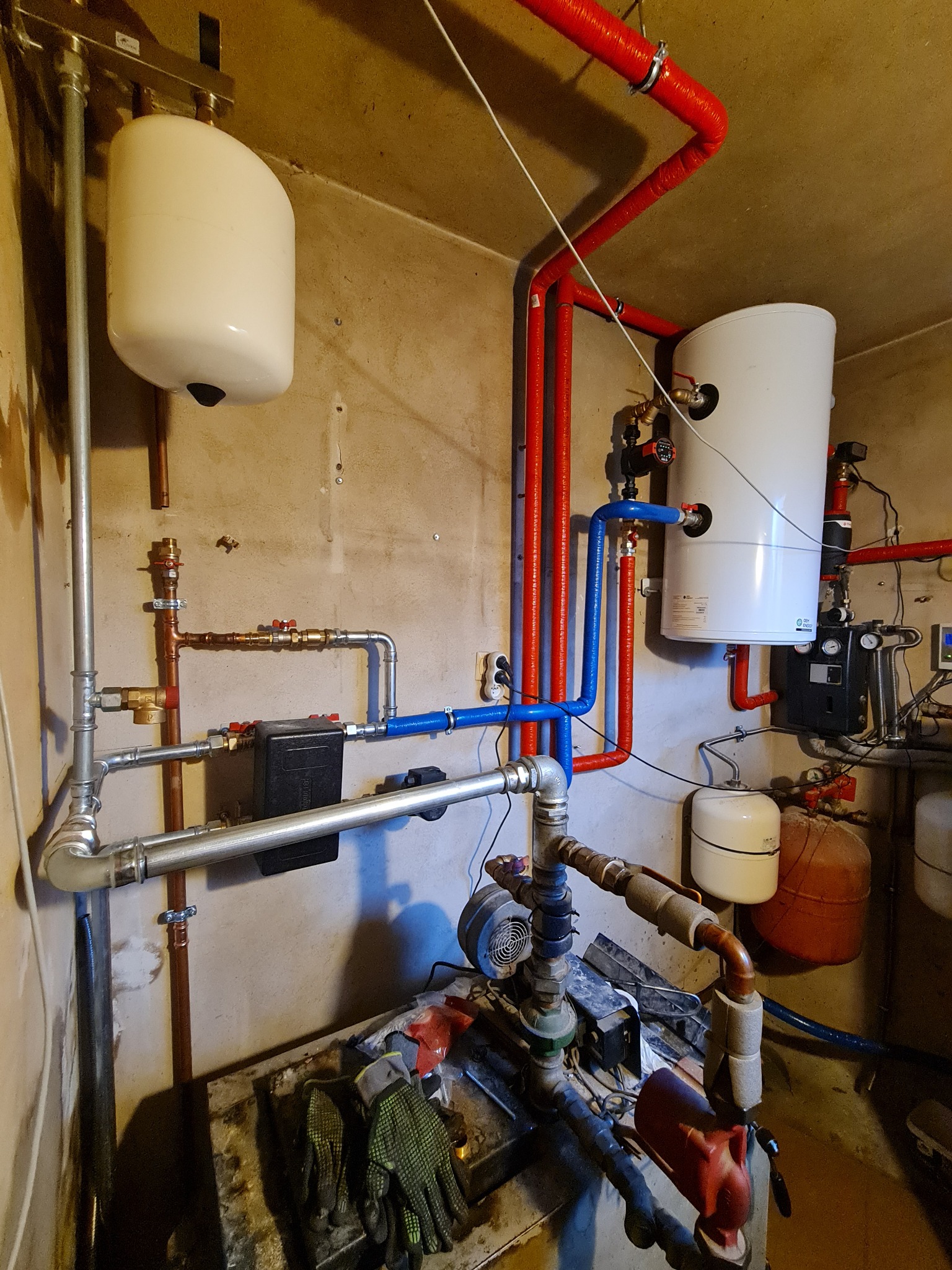 Złożona instalacja grzewcza w kotłowni: zbiorniki, rury miedziane i plastikowe w kolorach czerwonym i niebieskim, manometry, pompy obiegowe, widoczne rękawice robocze na podłodze.