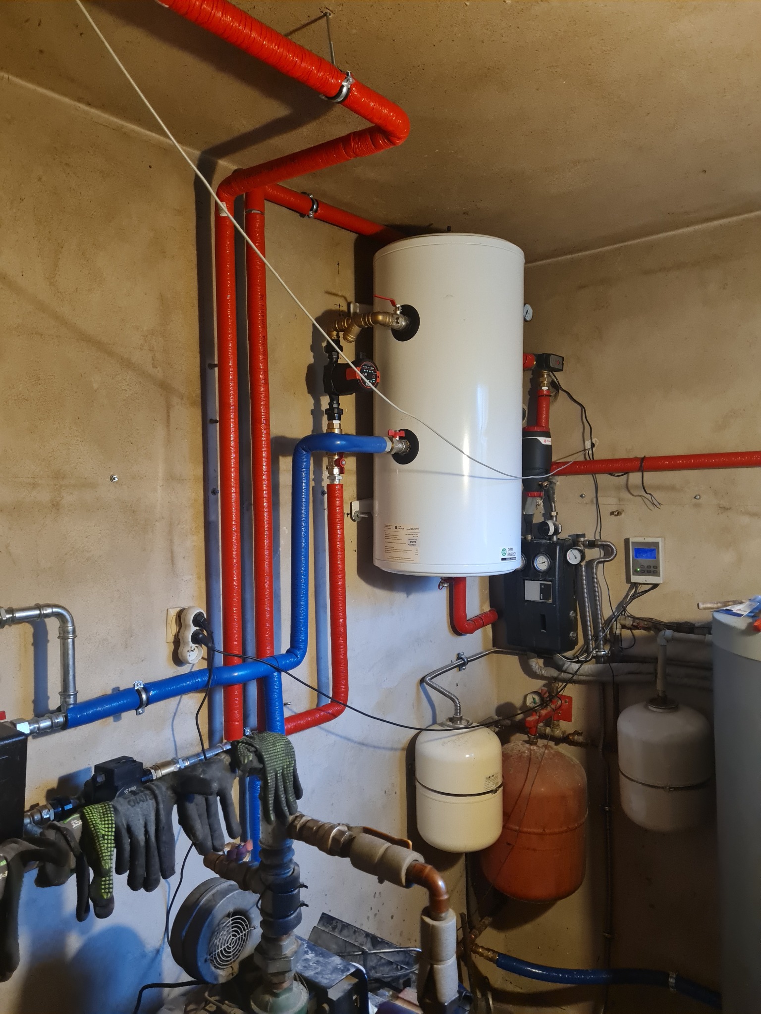 Instalacja pompy ciepła z widocznym zasobnikiem ciepłej wody, rurami w izolacji czerwonej i niebieskiej, oraz roboczymi rękawicami wiszącymi na rurach, sugerującymi trwające prace instalacyjne.