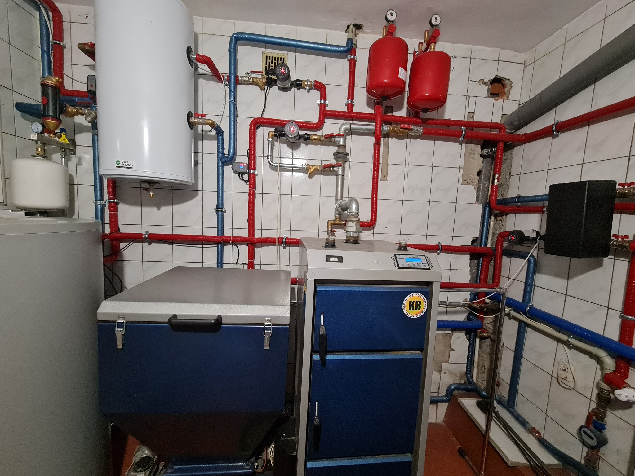 Kompletna instalacja centralnego ogrzewania z kotłem na paliwo stałe, zbiornikiem buforowym i rozbudowaną siecią rur miedzianych w izolacji, zamontowana w pomieszczeniu z białymi płytkami.