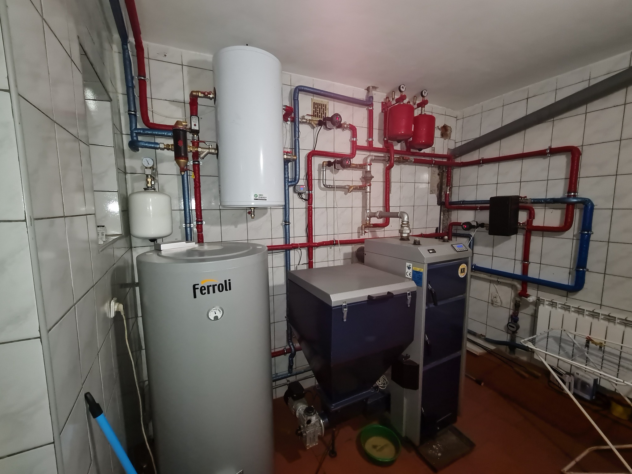 Kotłownia z zainstalowanym kotłem na paliwo stałe z podajnikiem, zbiornikiem buforowym Ferroli i rozbudowaną instalacją grzewczą z rurami w kolorach czerwonym i niebieskim.