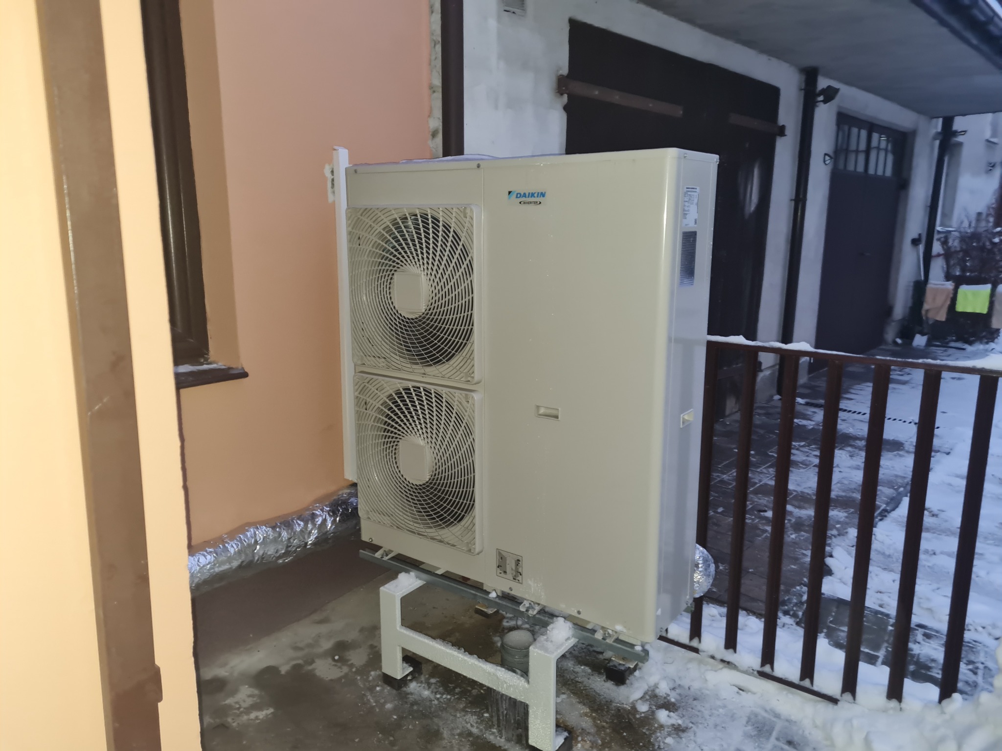Zewnętrzna jednostka klimatyzacji Daikin z dwoma wentylatorami, zamontowana na metalowym stelażu, na zewnątrz budynku w zimowej scenerii. Widoczna ściana budynku z oknem i fragment garażu w tle.