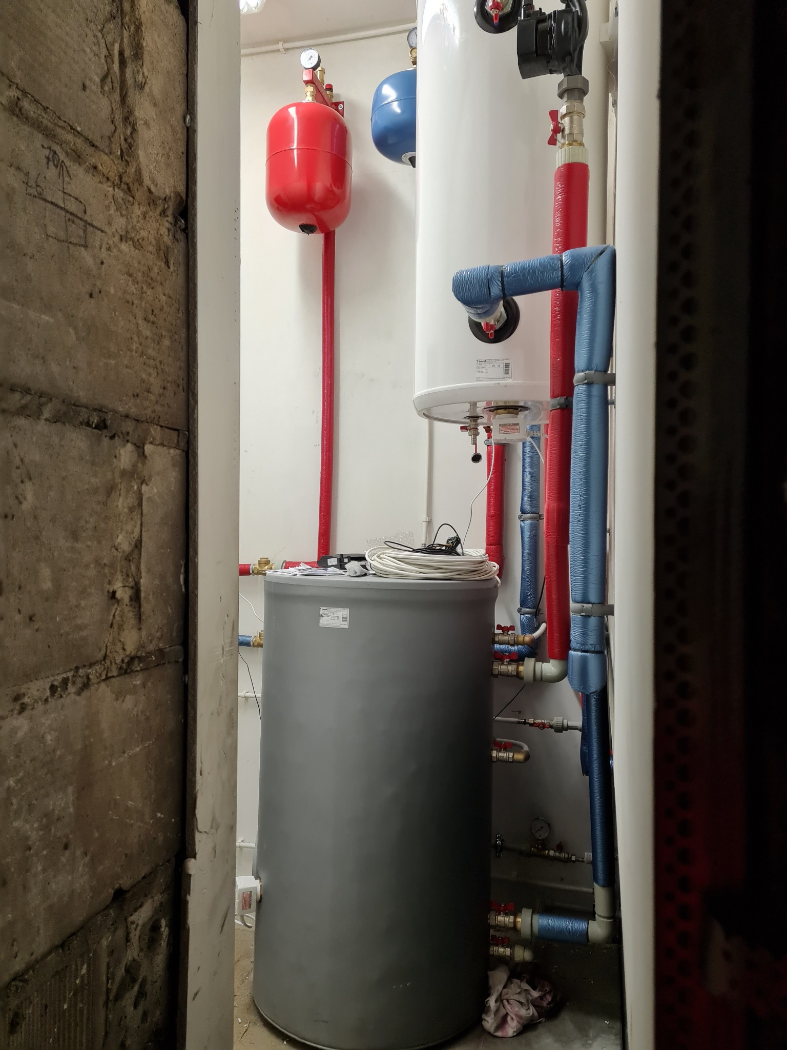 Instalacja centralnego ogrzewania z dwoma zbiornikami, czerwonym i niebieskim, oraz rurami w izolacji, widoczna w ciasnym pomieszczeniu z surową betonową ścianą po lewej stronie.
