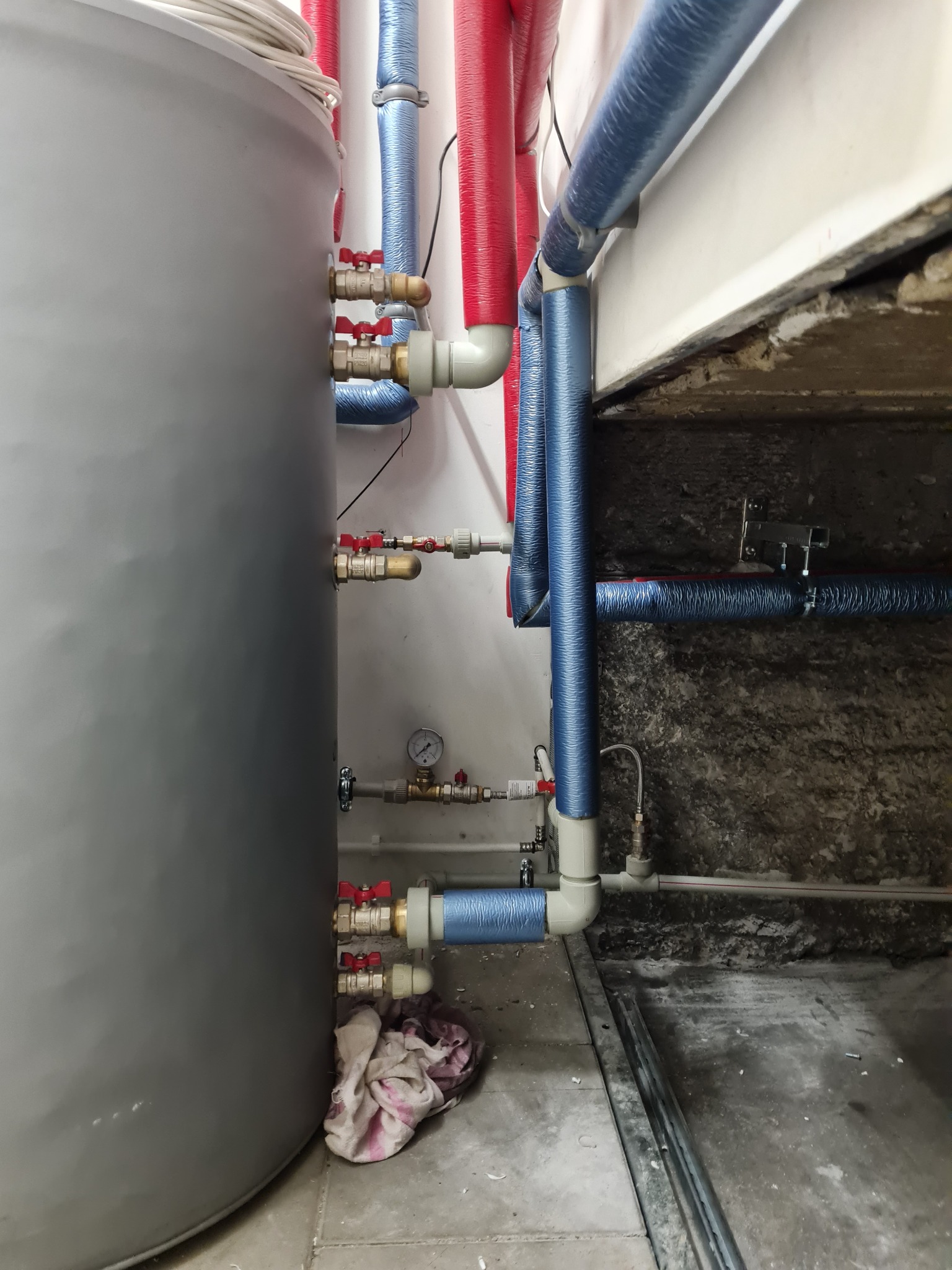 Szary zbiornik akumulacyjny z podłączonymi rurami w izolacji w kolorach czerwonym i niebieskim, widoczne zawory, manometr i ślady użytkowania na podłodze.