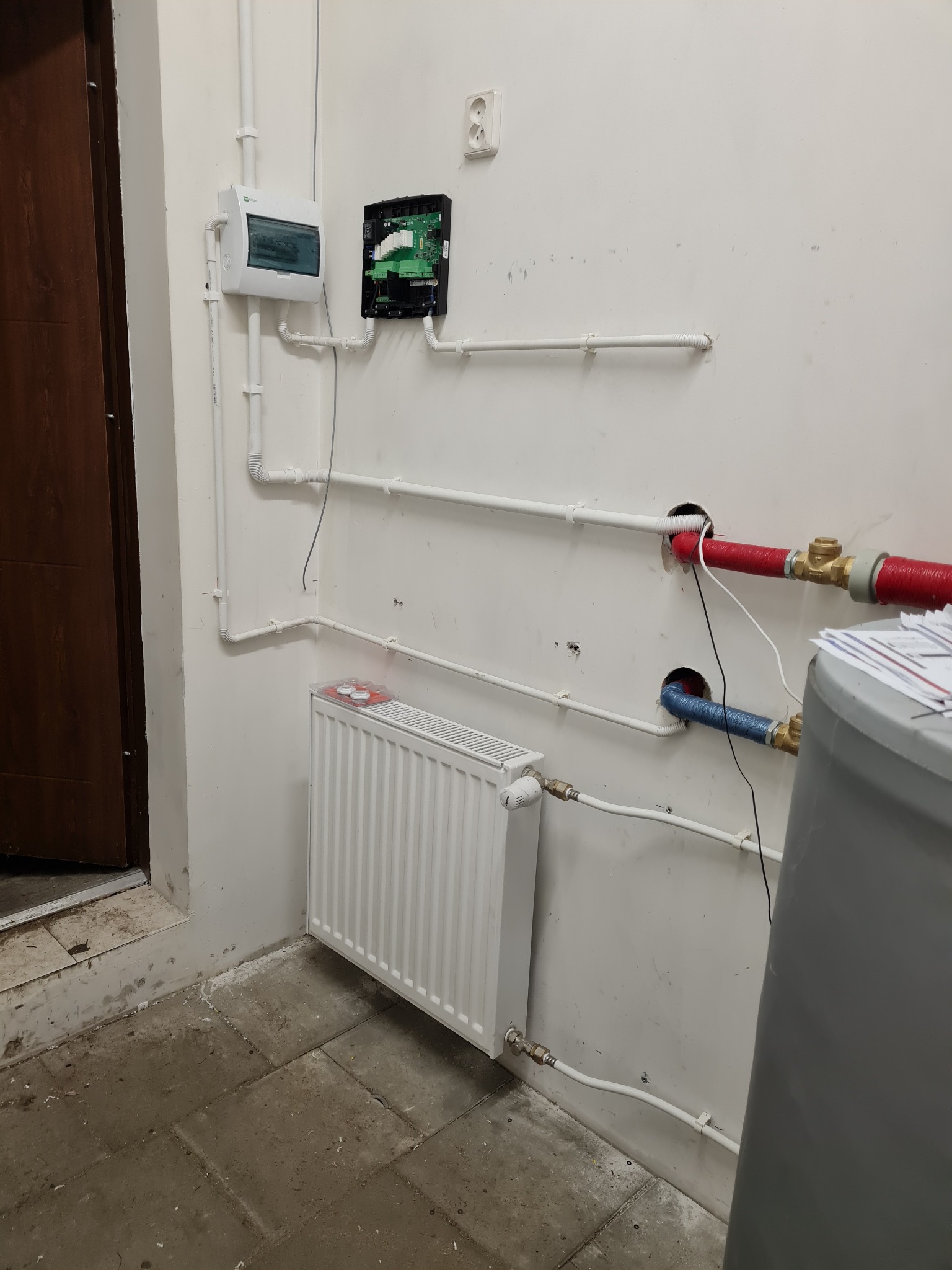 Instalacja centralnego ogrzewania z grzejnikiem panelowym, zbiornikiem i przewodami w pomieszczeniu technicznym, widoczne elementy sterujące na ścianie.