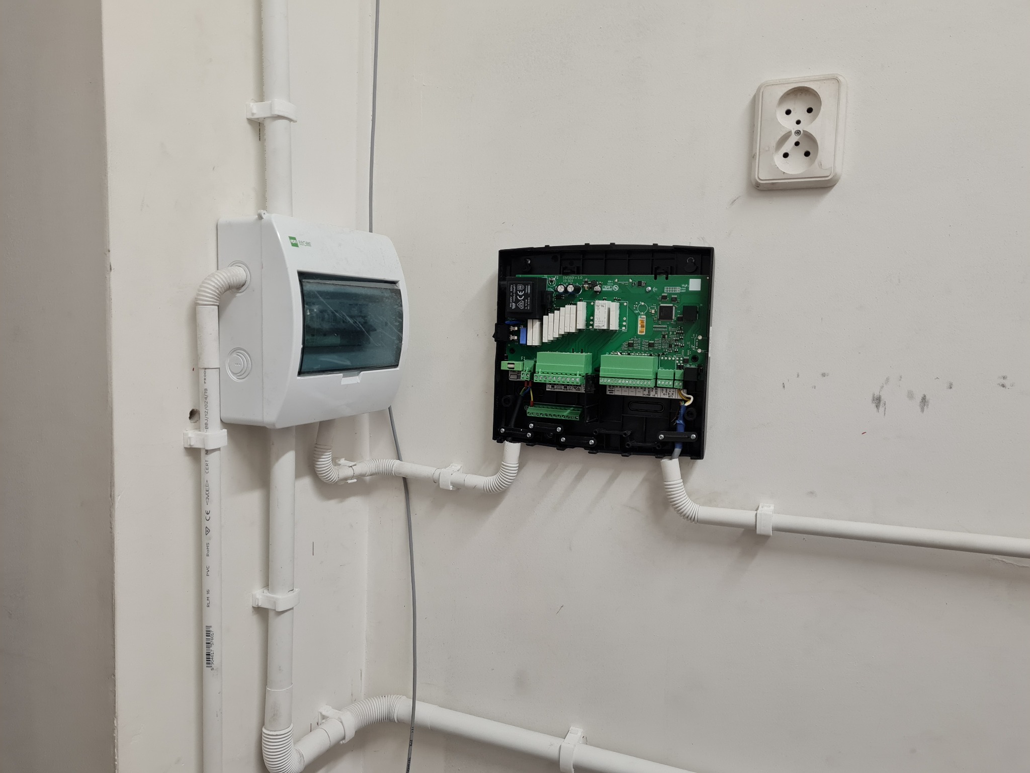 Rozdzielnia elektryczna i sterownik pompy ciepła zamontowane na białej ścianie, widoczne rury instalacyjne i gniazdko elektryczne