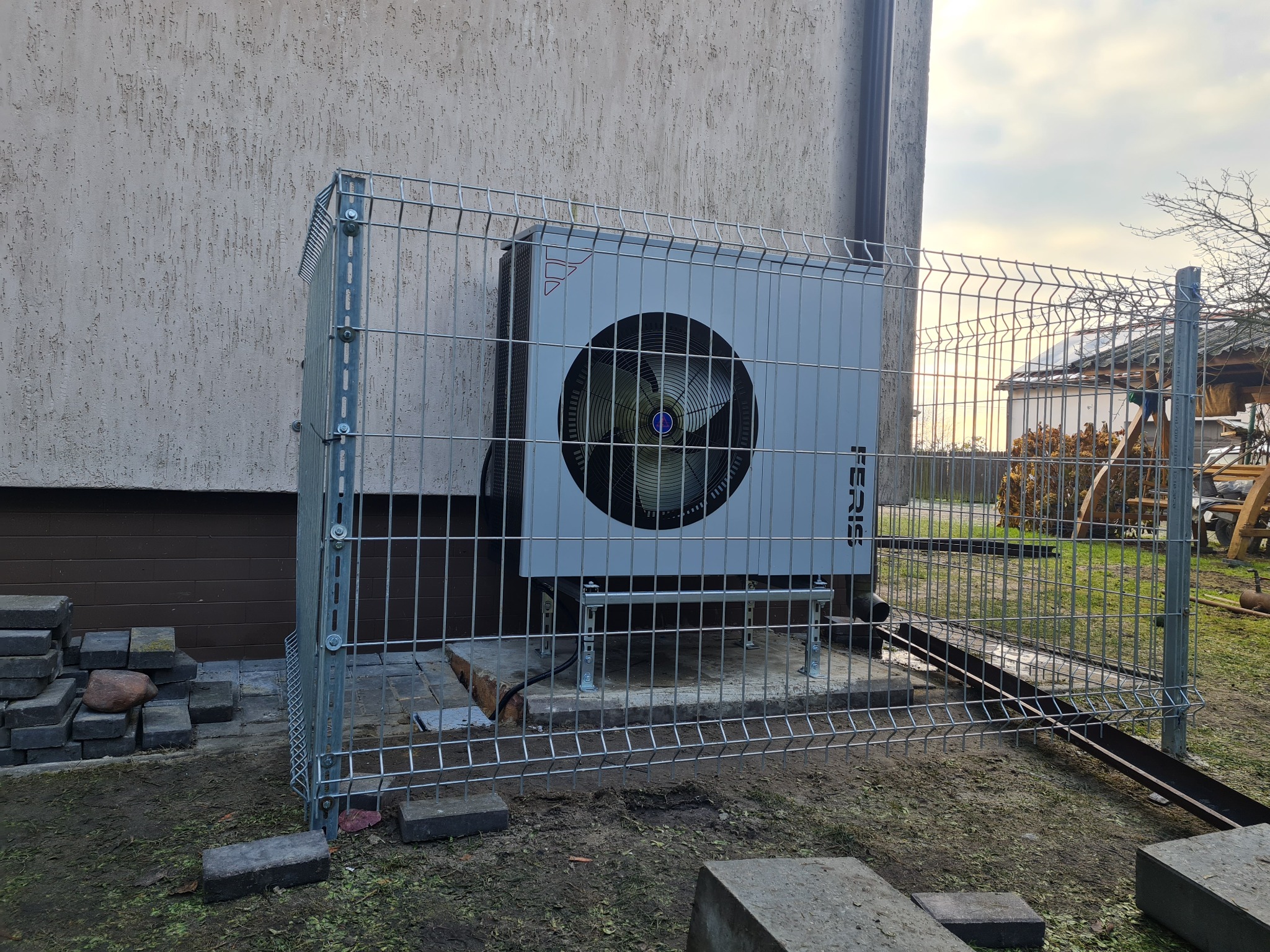 Szara pompa ciepła marki Feris zamontowana na betonowej podstawie, zabezpieczona metalową klatką w ogrodzie, widok zza ogrodzenia.