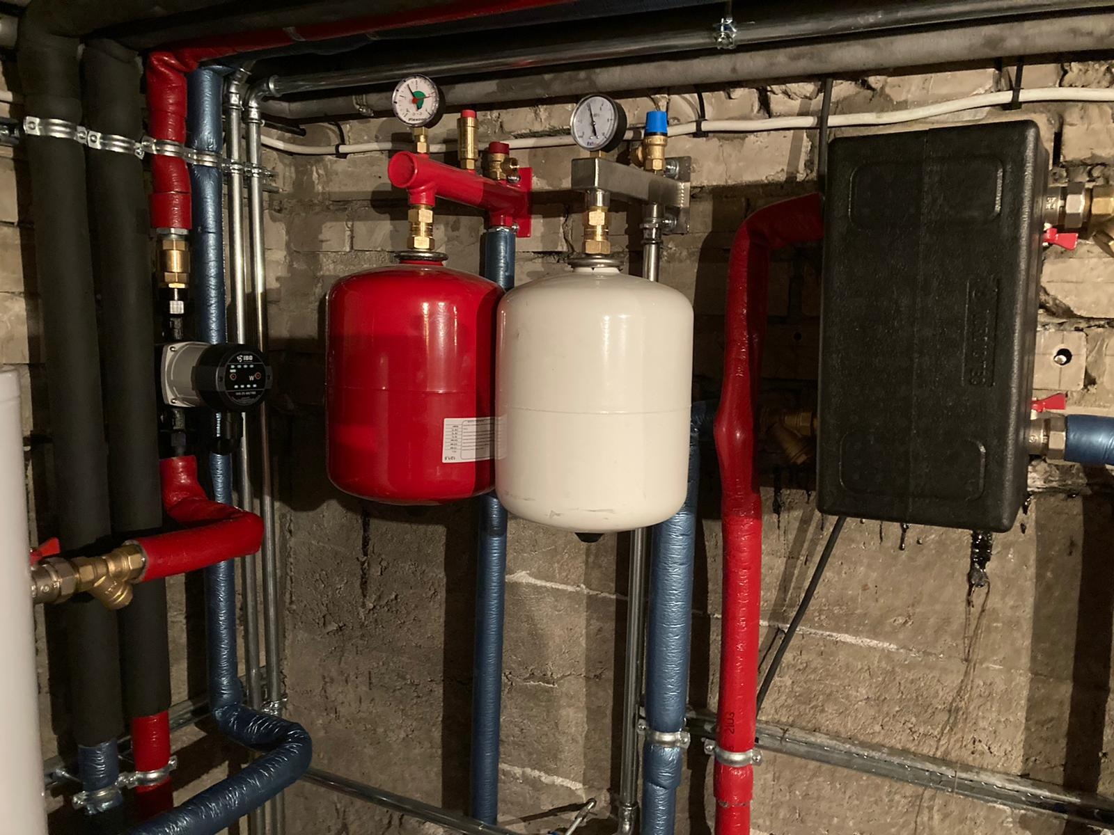 Instalacja centralnego ogrzewania z czerwonym i białym zbiornikiem wyrównawczym, manometrami i rurami w izolacji na tle surowej ściany.