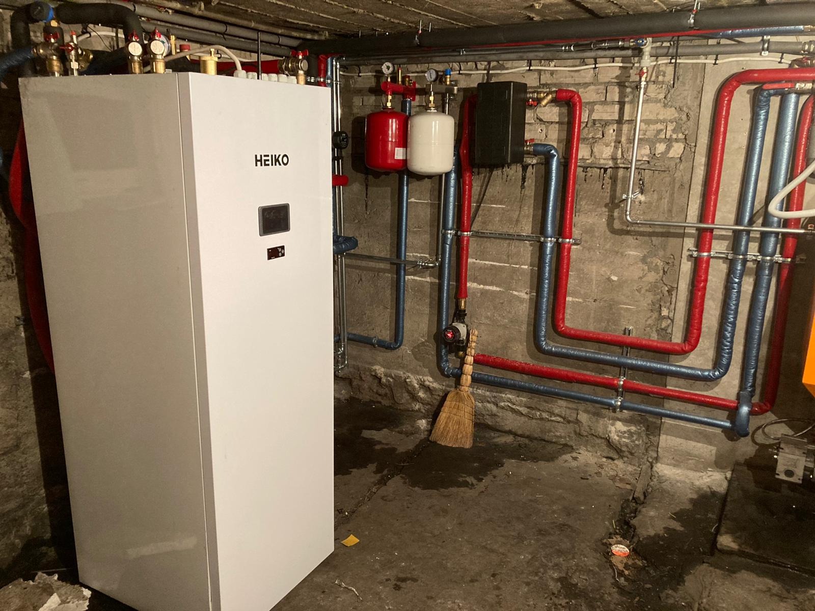 Biała pompa ciepła marki HEIKO w piwnicy, widoczne rury w izolacji termicznej, czerwone i niebieskie, oraz stara ceglana ściana. Na podłodze stoi miotła.