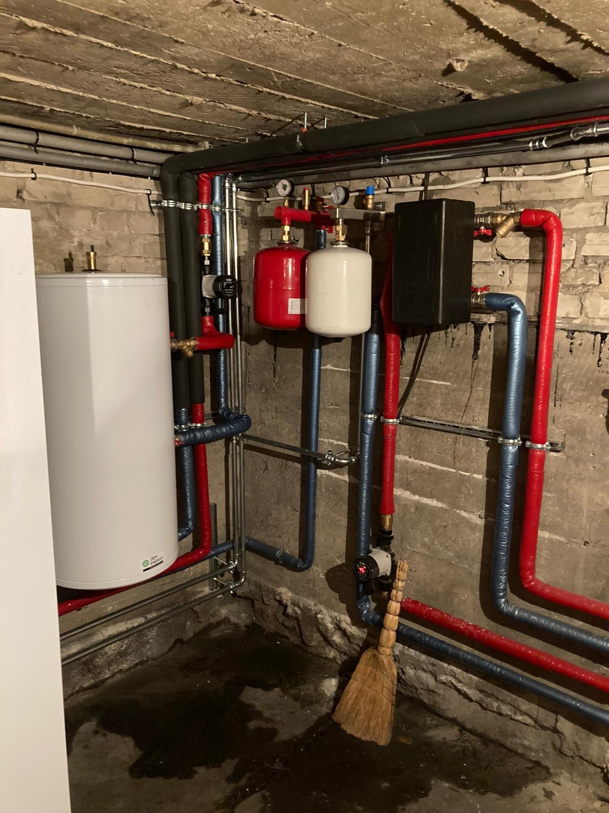 Instalacja pompy ciepła w piwnicy: biały zbiornik buforowy, czerwone i niebieskie rury, czarny sterownik, tło z surowego betonu, obok stoi miotła.