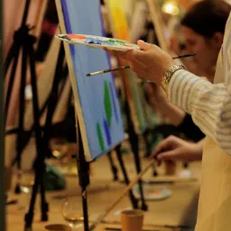 Uczestnicy malują obrazy na pł&oacute;tnach podczas warsztat&oacute;w artystycznych, widoczne sztalugi, pędzle, palety z farbami oraz napoje.