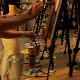 Uczestnicy warsztatów malarskich przy sztalugach, malujący obrazy na płótnach, z paletami farb i pędzlami na stole.