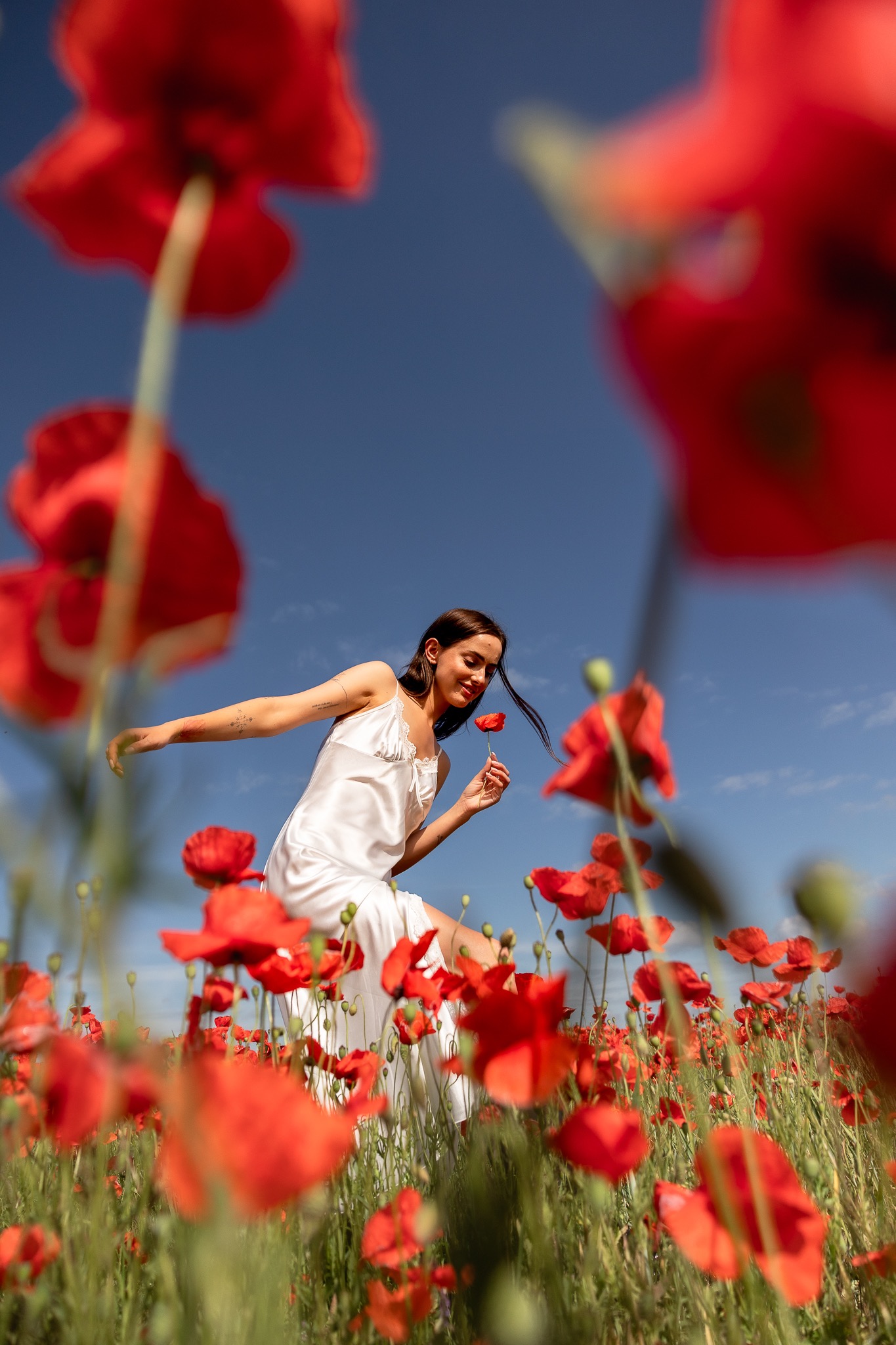 Kobieta w białej sukience stoi na polu maków, trzymając jeden kwiat w dłoni, błękitne niebo w tle.