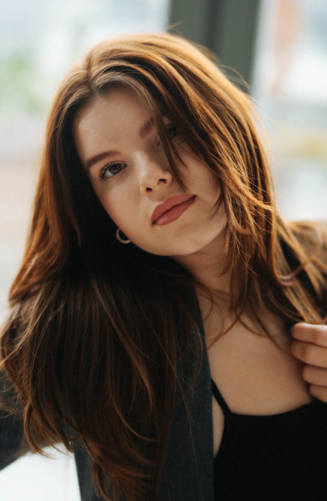 Portret młodej kobiety z długimi, rudymi włosami, delikatny makijaż, skierowana w dół, ujęcie z góry, subtelne oświetlenie okienne.