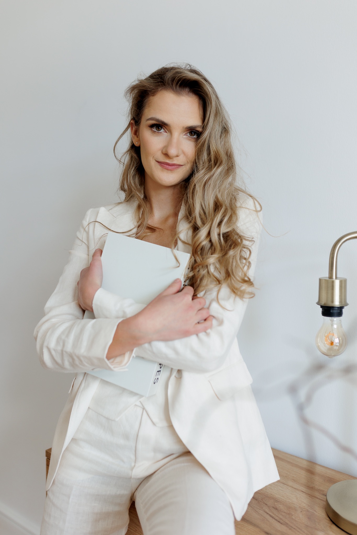 Uśmiechnięta kobieta w białym garniturze, trzymająca jasną teczkę z napisem, pozująca przy biurku z lampką w stylu retro.