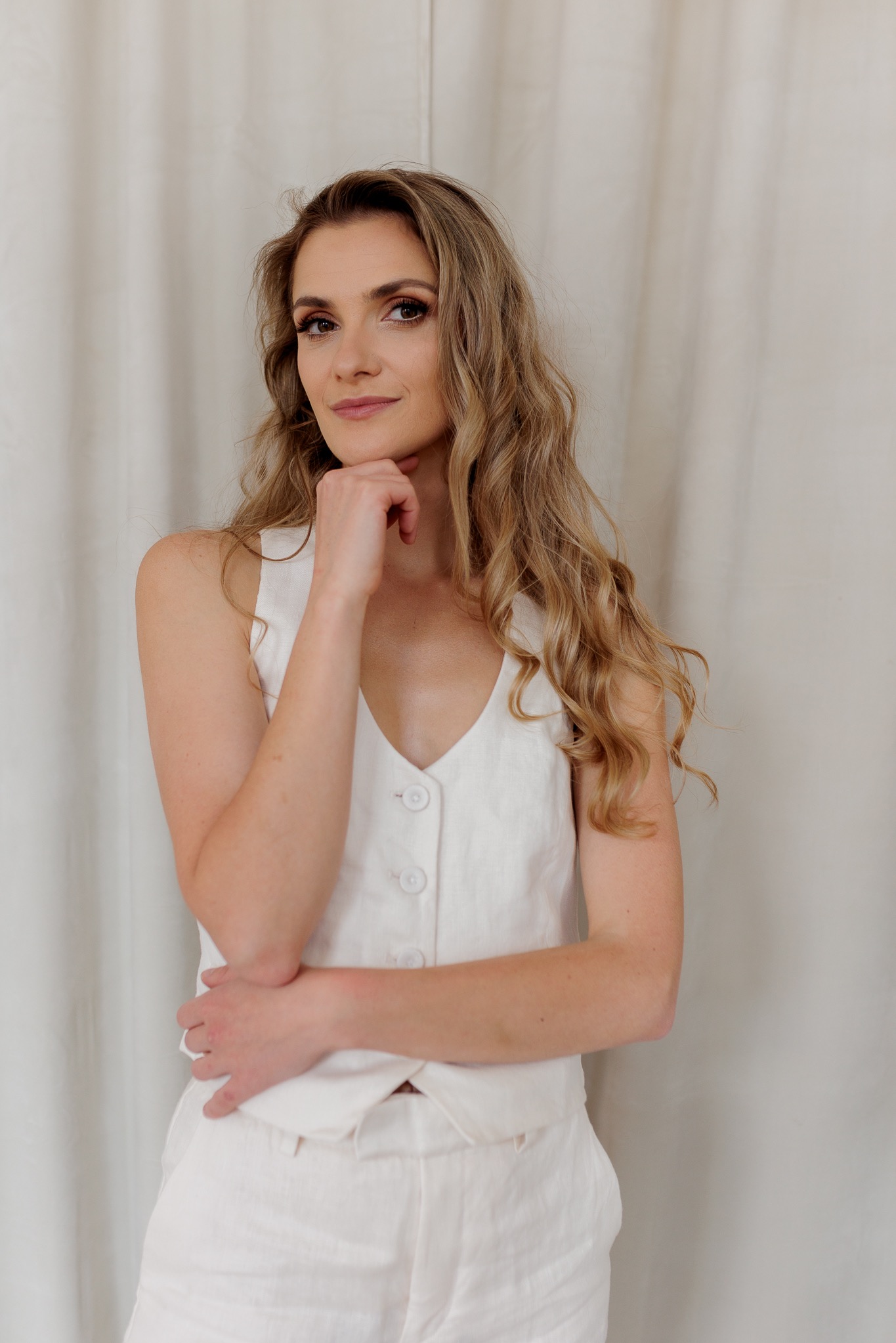 Portret kobiety z długimi blond włosami, ubrana w biały lniany komplet, pozująca z ręką pod brodą na tle kremowej zasłony.