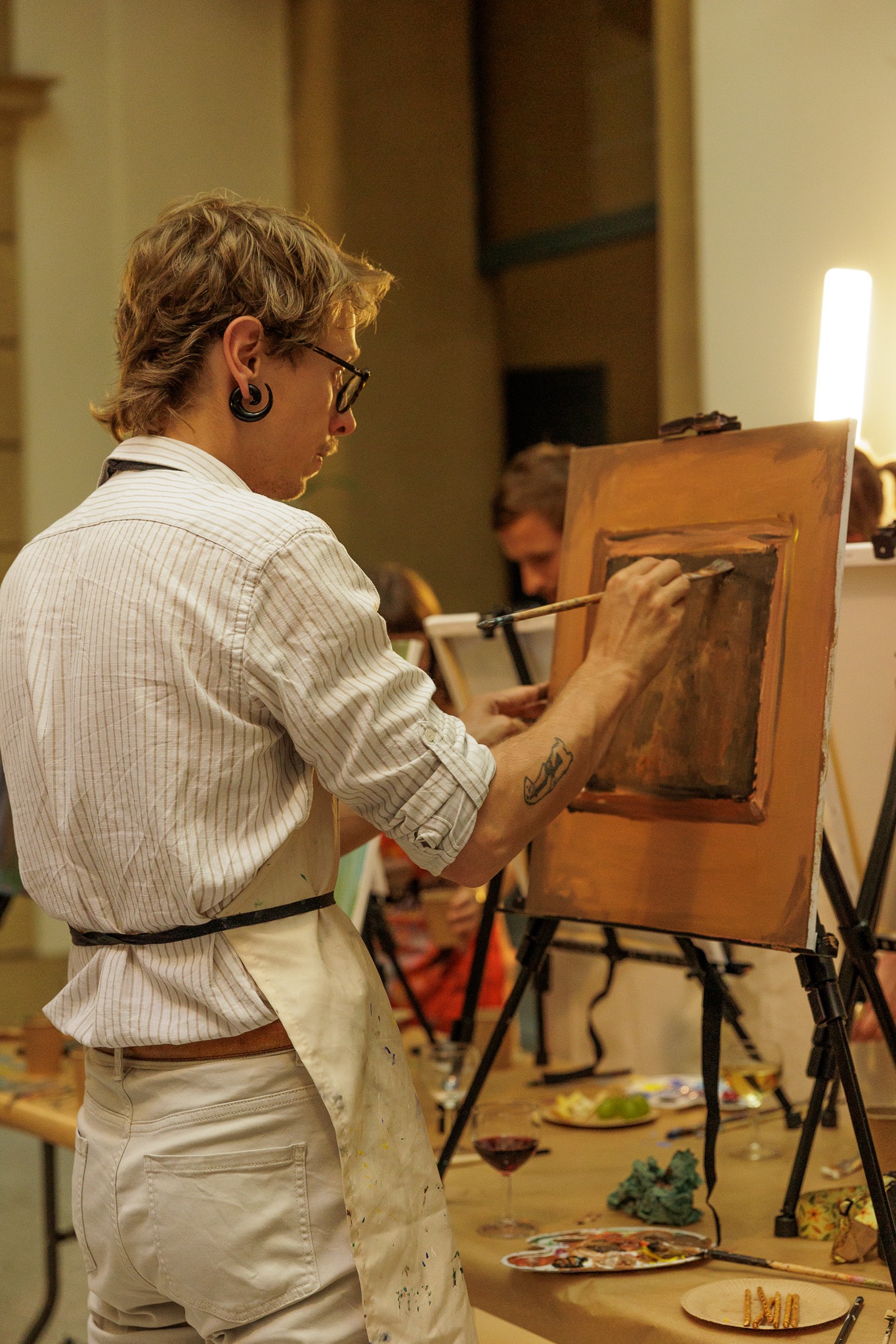 Artysta z tunelem w uchu maluje obraz na płótnie podczas warsztatów malarskich; widoczny kieliszek wina i paleta z farbami.