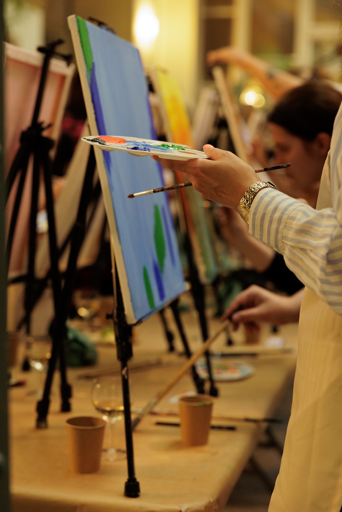 Uczestnicy malują obrazy na płótnach podczas warsztatów artystycznych, widoczne sztalugi, pędzle, palety z farbami oraz napoje.