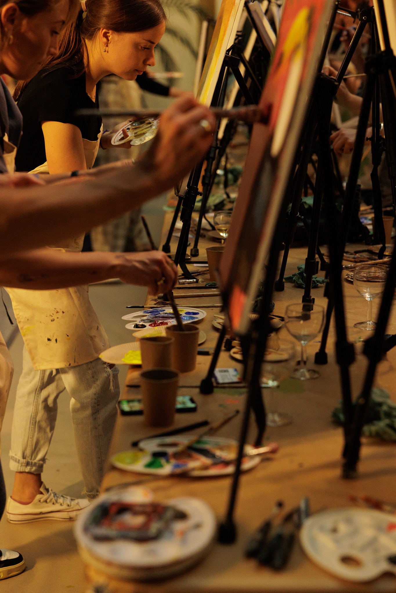 Uczestnicy warsztatów malarskich przy sztalugach, malujący obrazy na płótnach, z paletami farb i pędzlami na stole.