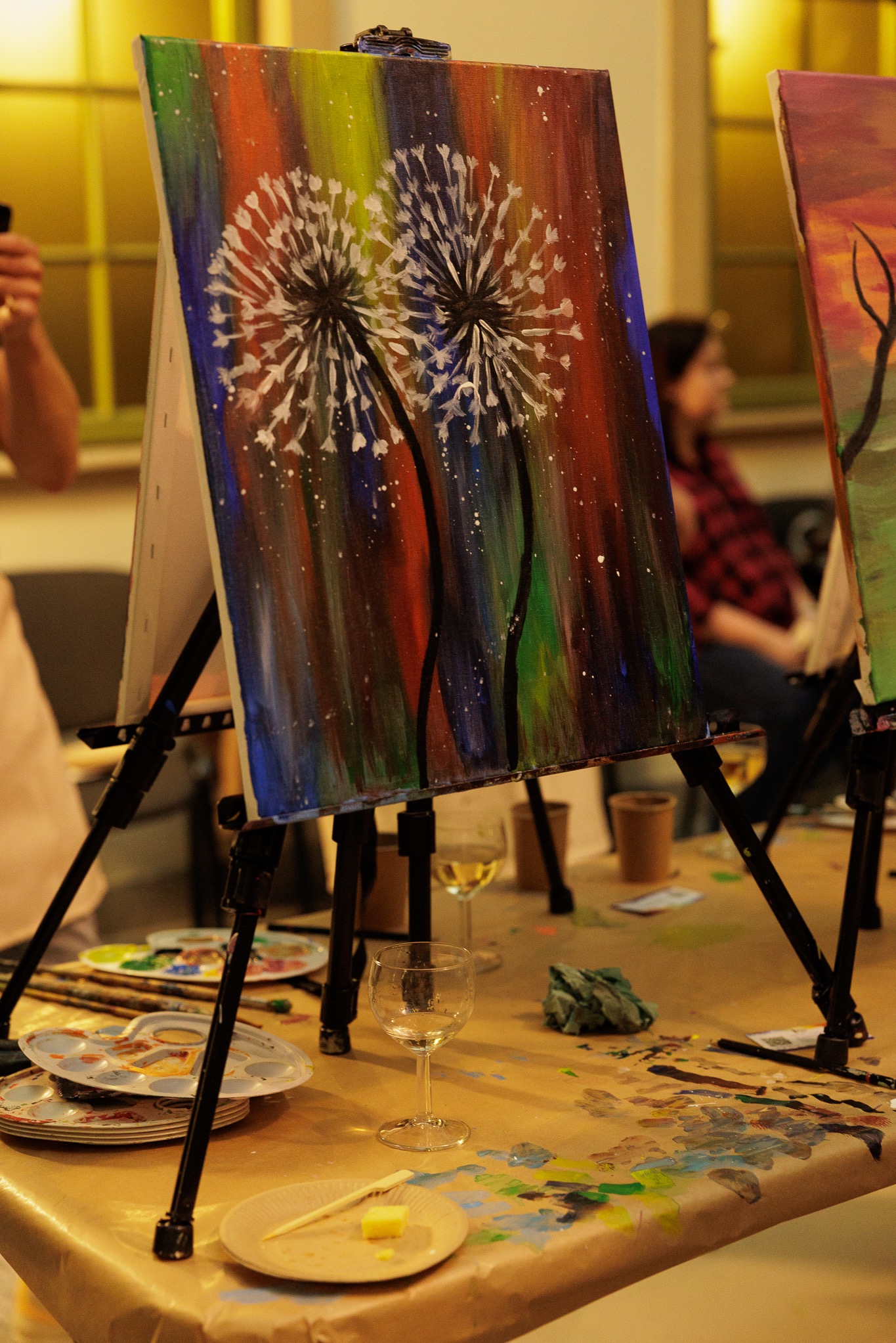 Dwa obrazy z motywem dmuchawców na sztalugach, malowane na płótnie, z kieliszkiem wina i paletami farb na stole, w tle siedząca osoba w czerwonej koszuli w kratę.