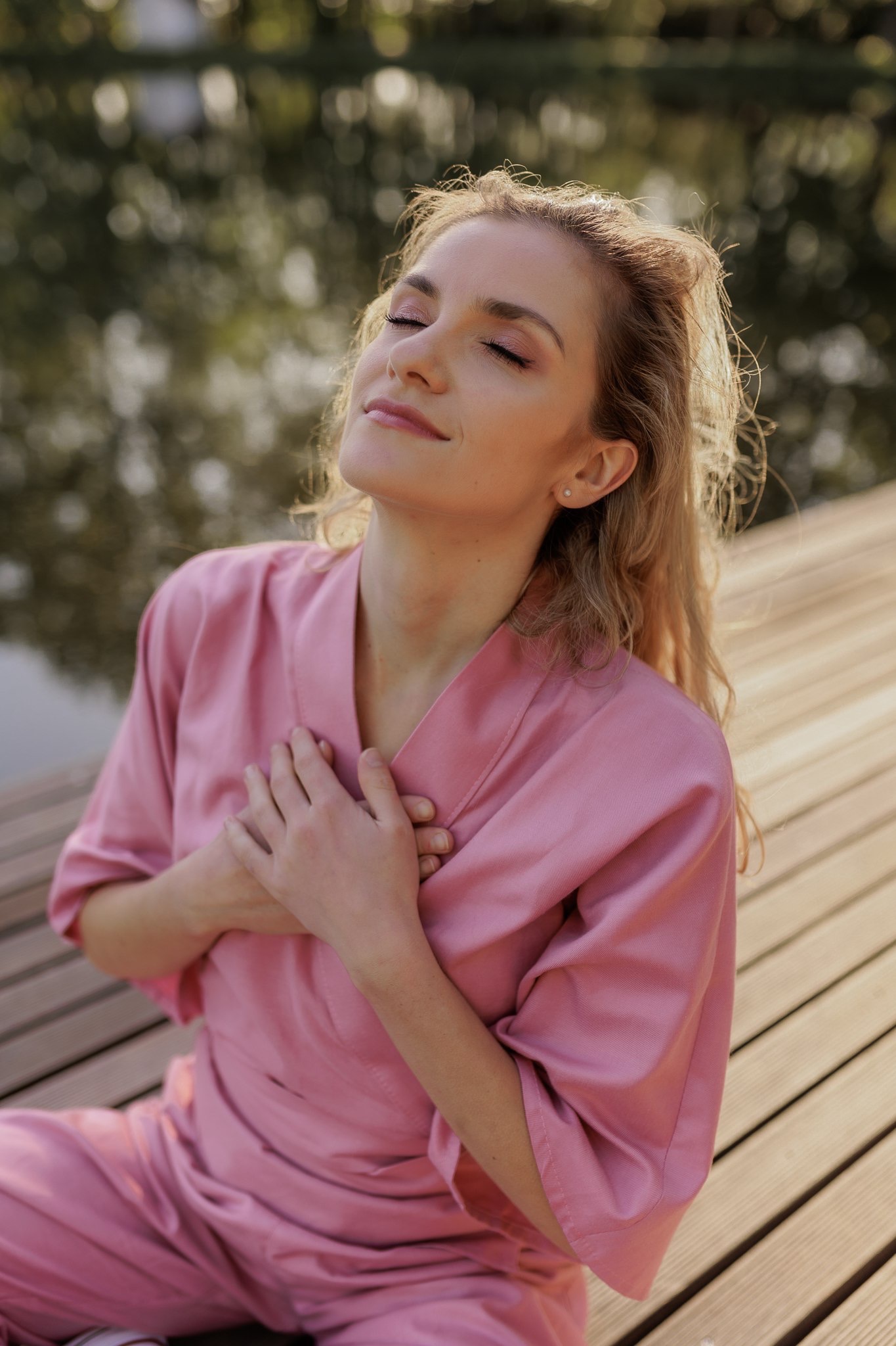 Portret kobiety w różowym szlafroku siedzącej na drewnianym pomoście z rękami na piersi, z zamkniętymi oczami i rozproszonym światłem w tle.