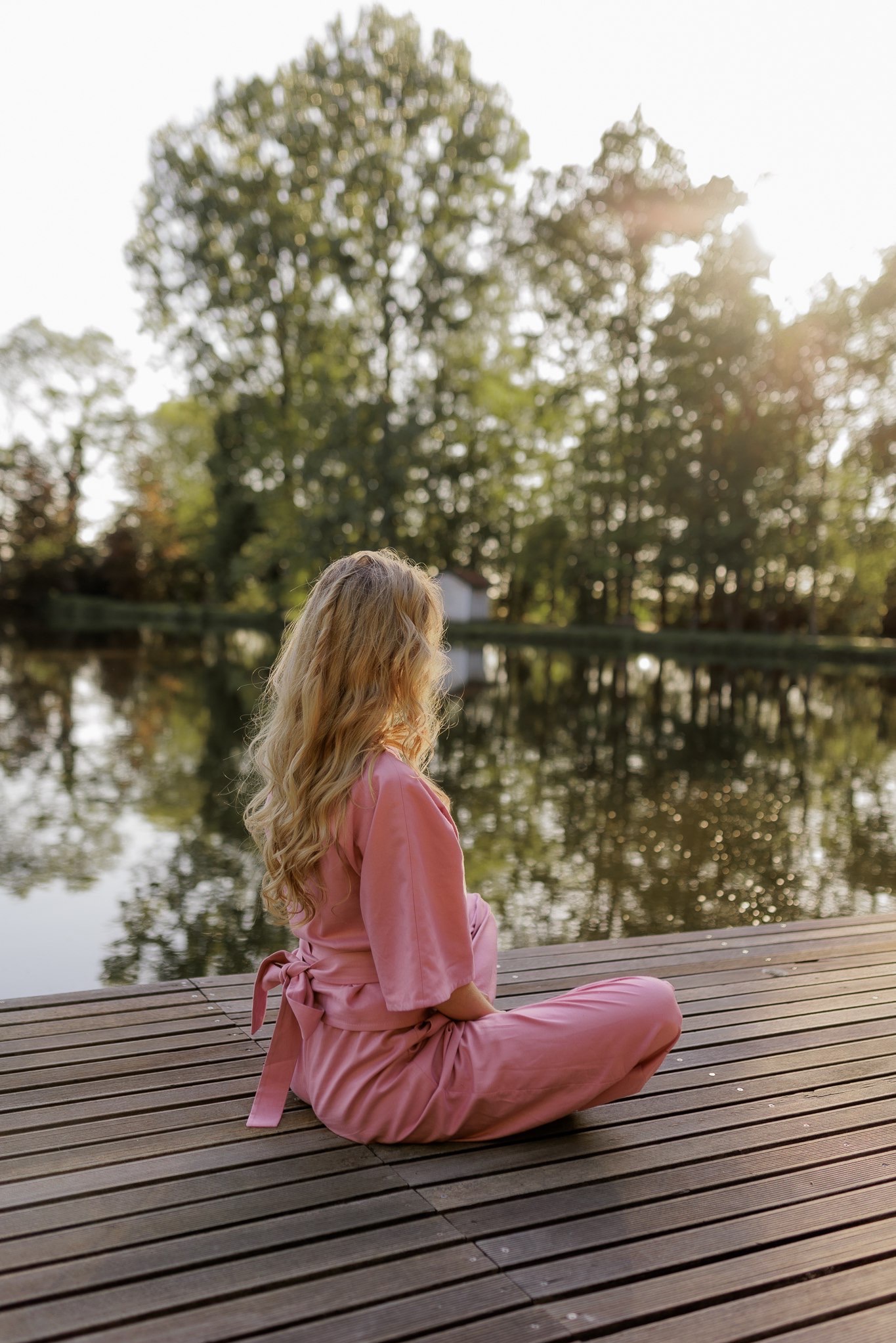 Kobieta w różowym stroju siedzi tyłem na drewnianym pomoście nad jeziorem, z widokiem na drzewa i odbicia w wodzie.
