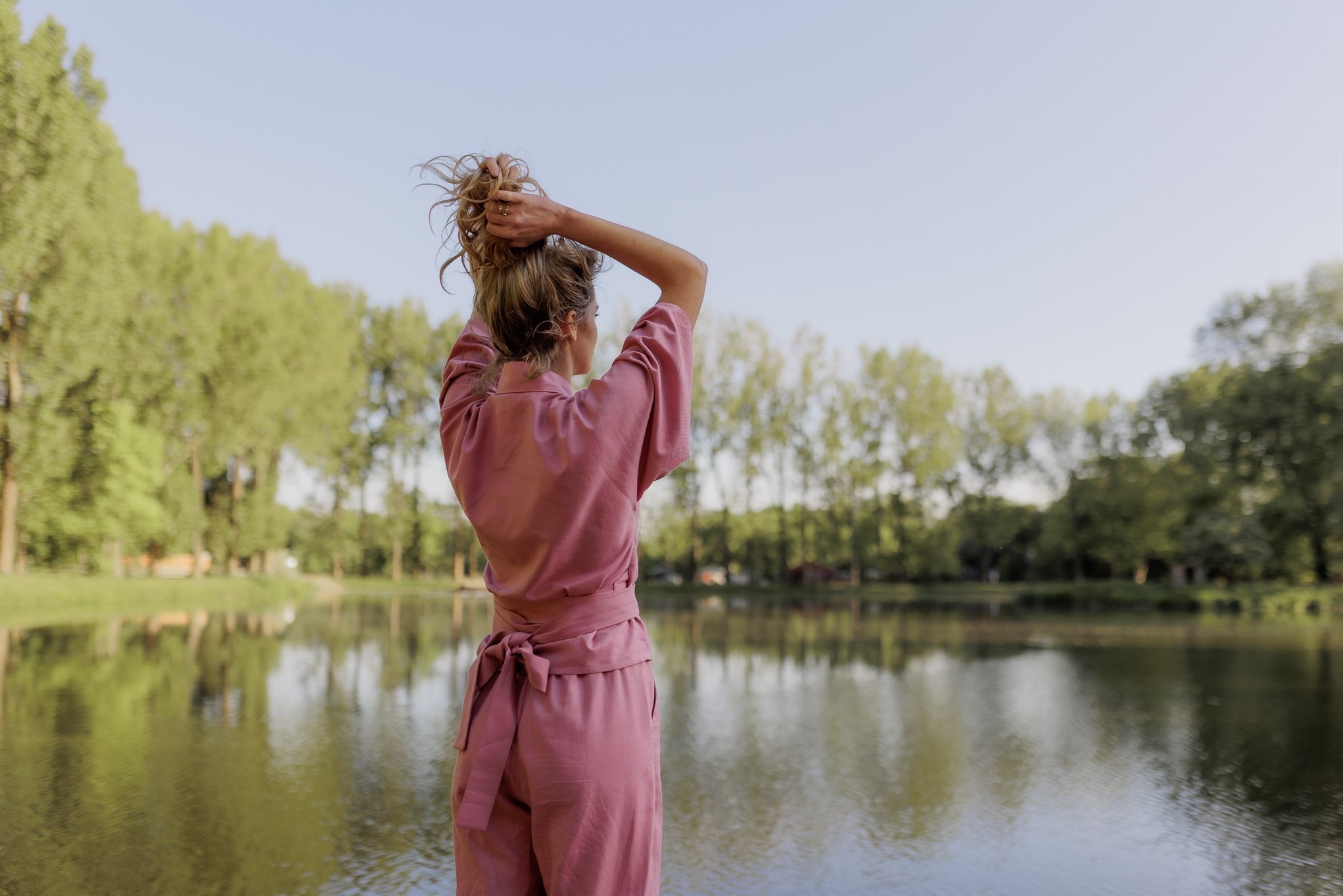 Kobieta w różowym kombinezonie, stojąca tyłem nad brzegiem jeziora, podnosi ręce do włosów, tworząc luźny kok. W tle zielone drzewa i błękitne niebo.