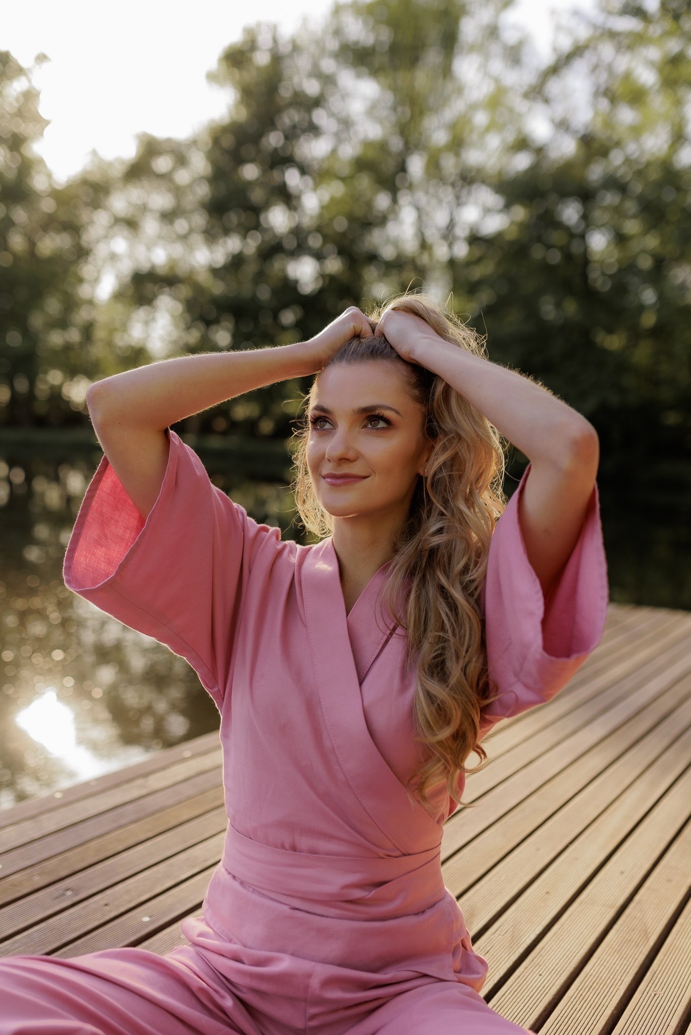 Portret kobiety w różowym kimonie siedzącej na drewnianym pomoście nad wodą, z rękami uniesionymi do włosów i zamyślonym spojrzeniem w górę.