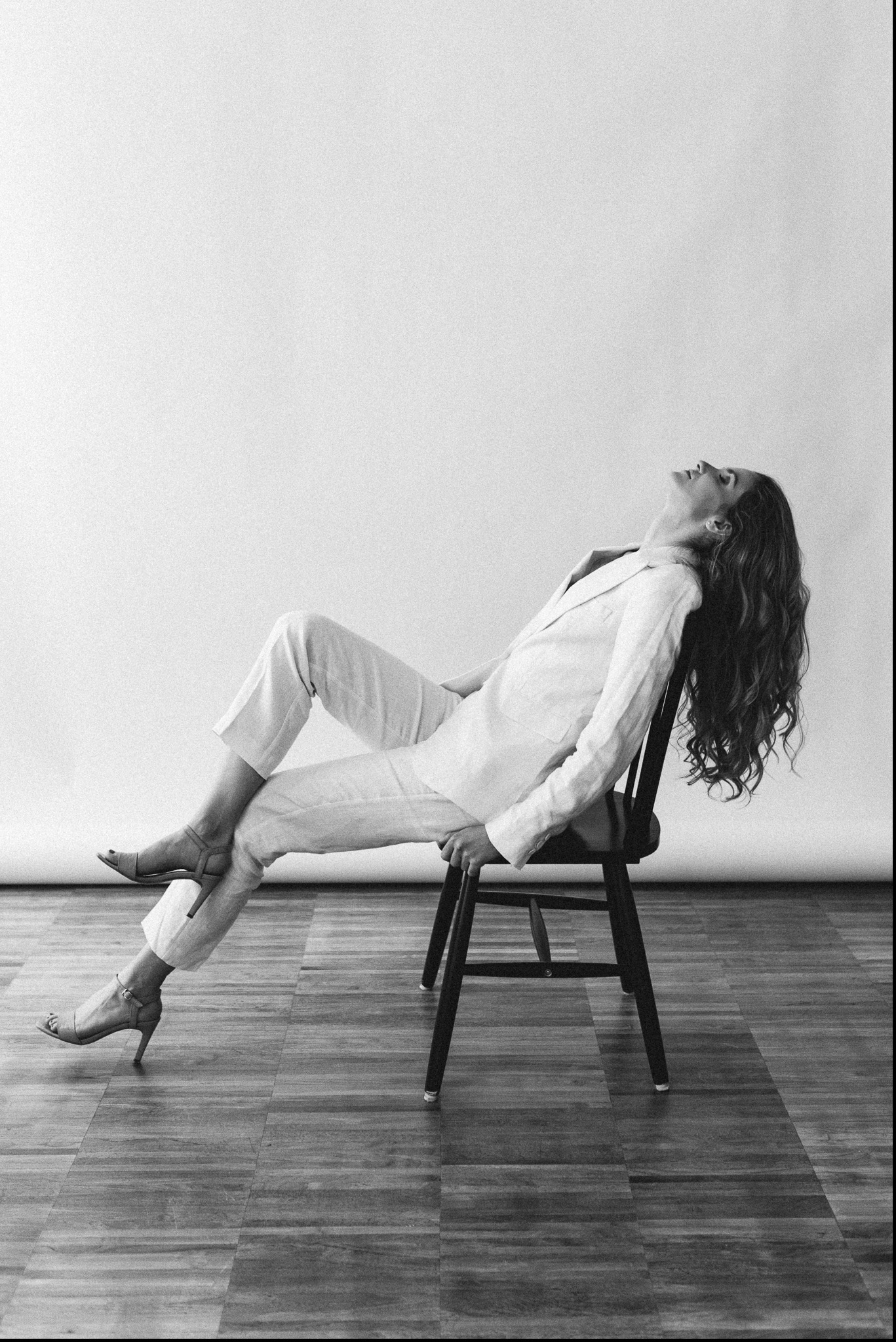 Kobieta w białym garniturze i szpilkach leży odchylona na krześle, głowa do góry, na tle jasnej ściany i drewnianej podłogi.