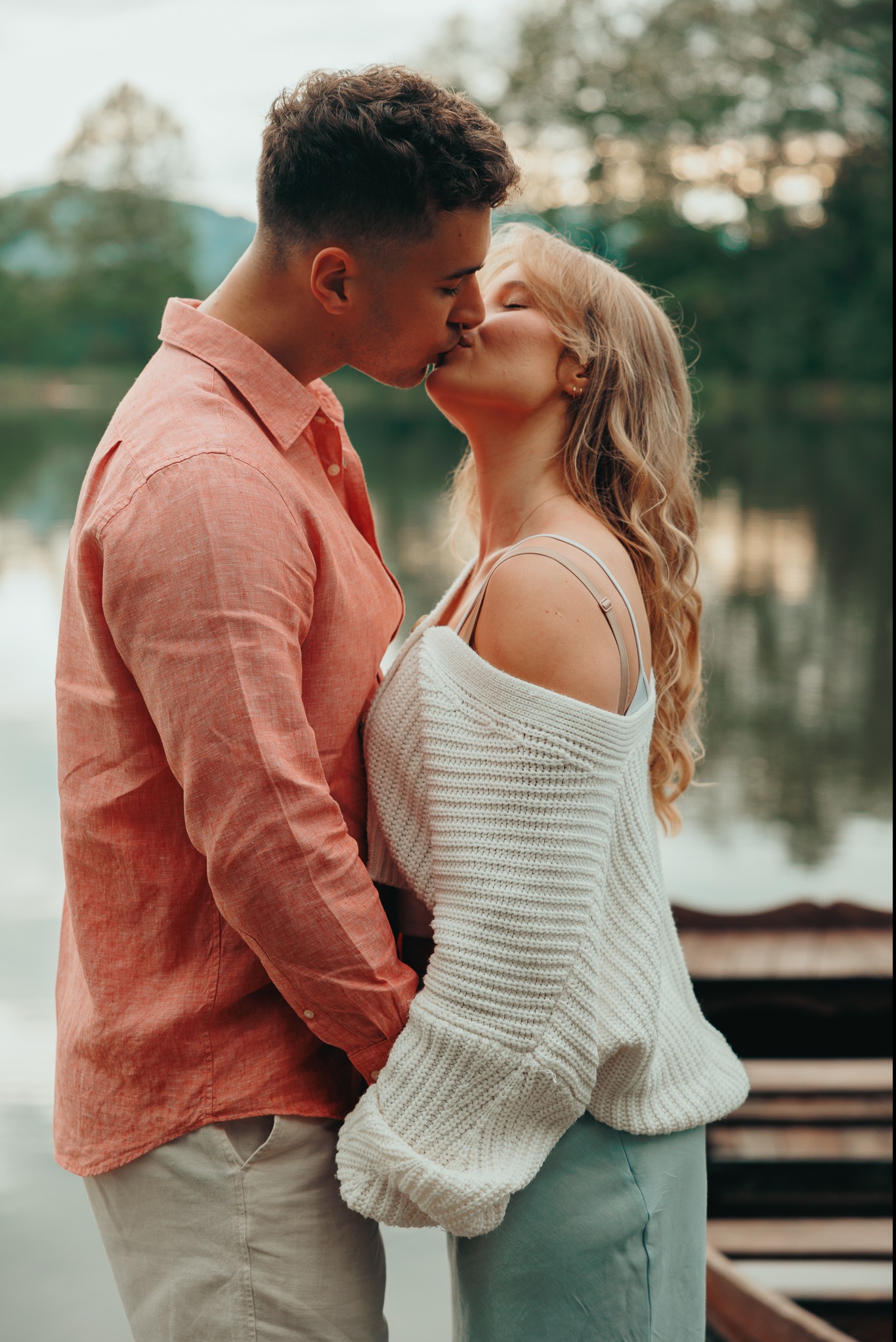 Zakochana para całuje się na tle jeziora, ona w białym swetrze i zielonej spódnicy, on w pomarańczowej koszuli i beżowych spodniach.