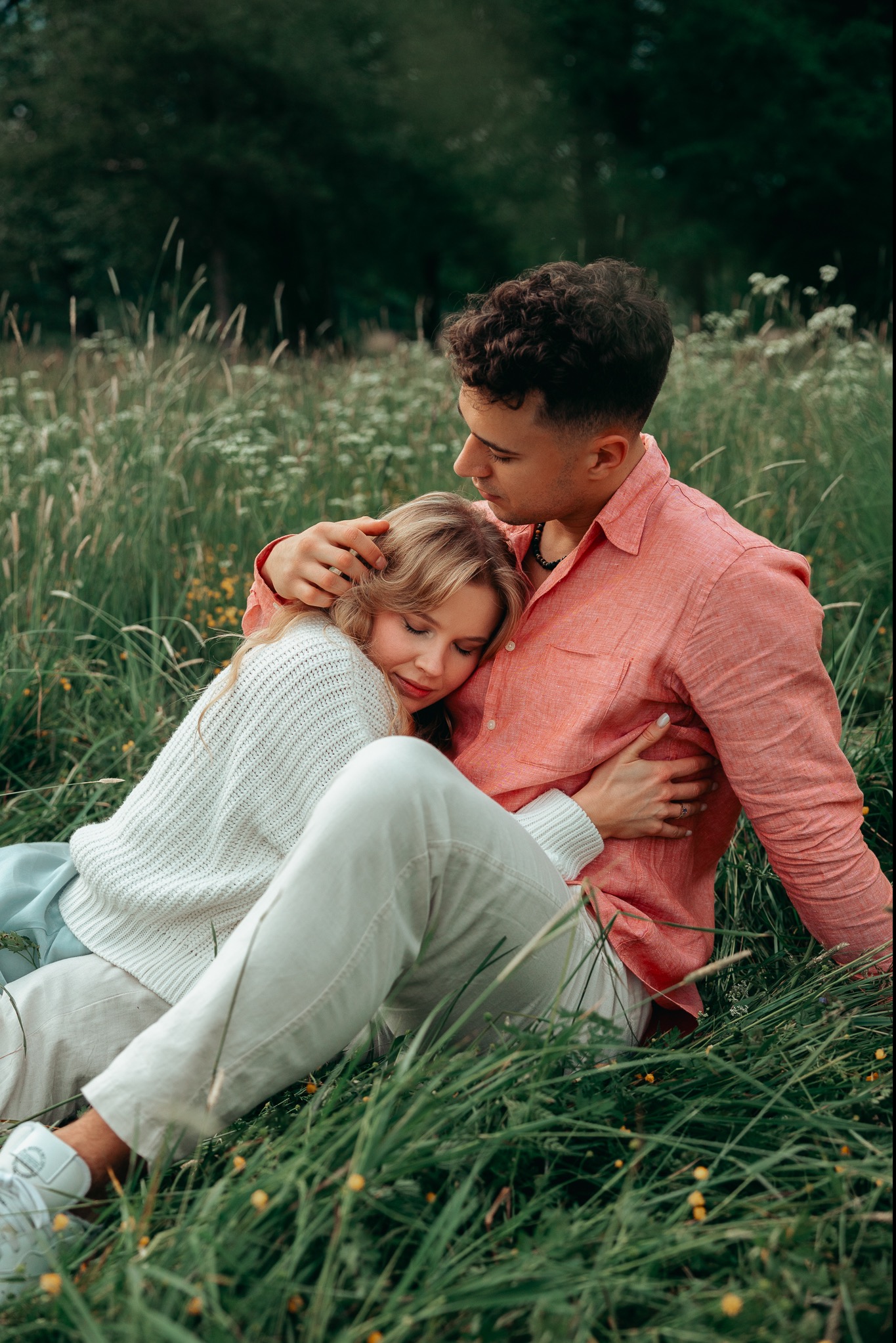 Młoda para: mężczyzna w koralowej koszuli obejmuje kobietę w białym swetrze, siedzącą na trawie z zamkniętymi oczami.