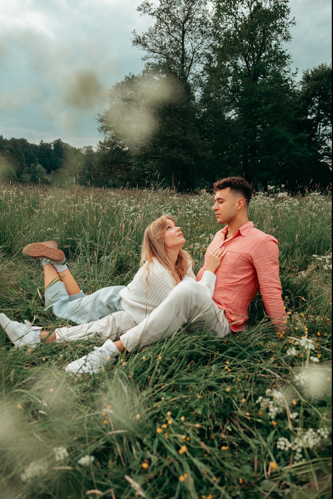 Para leżąca na łące w wysokiej trawie, kobieta w białym swetrze spogląda na mężczyznę w różowej koszuli, tło z drzewami i pochmurnym niebem.