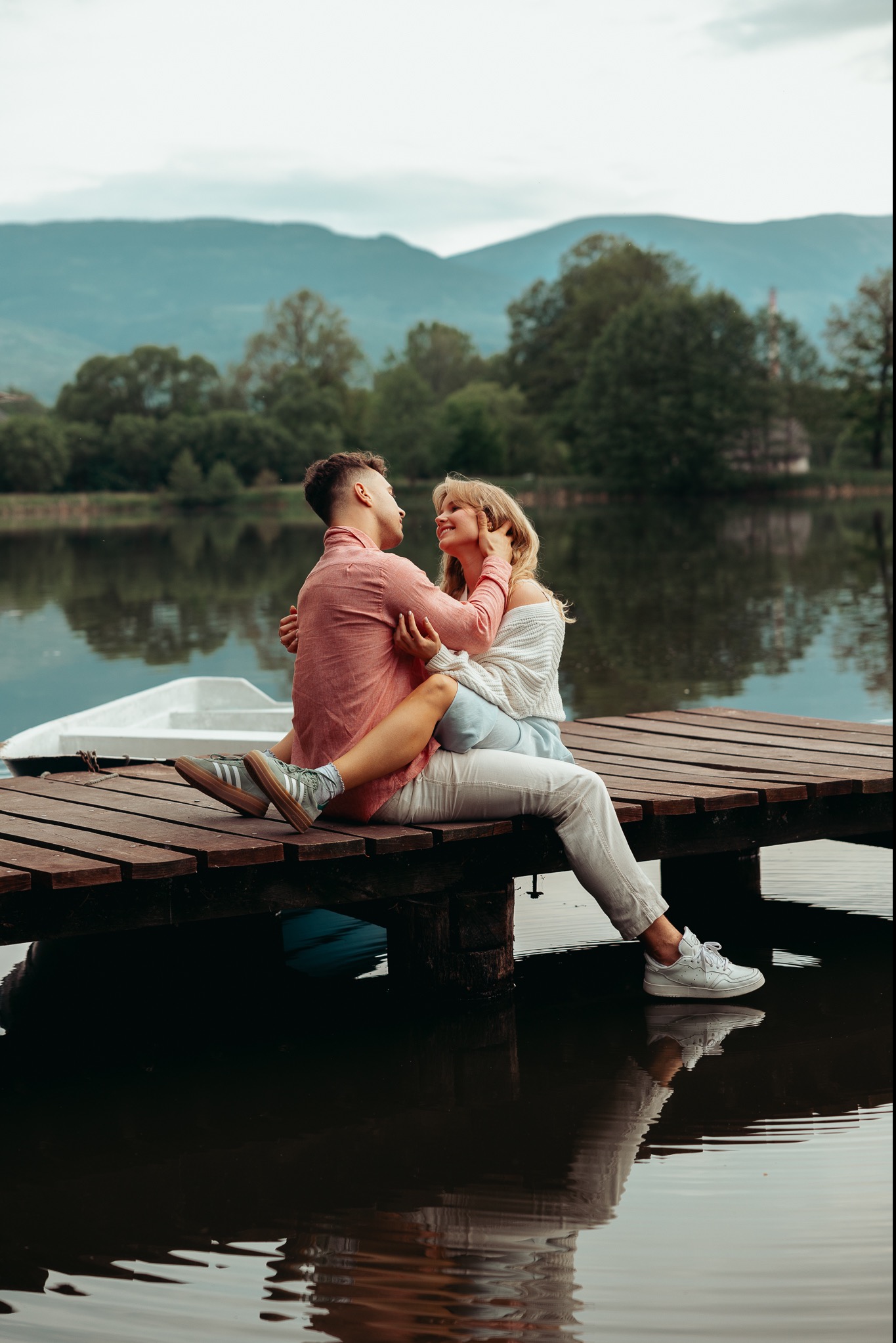 Para siedząca na drewnianym pomoście nad jeziorem, mężczyzna w różowej koszuli obejmuje kobietę w białym swetrze, w tle góry i drzewa.