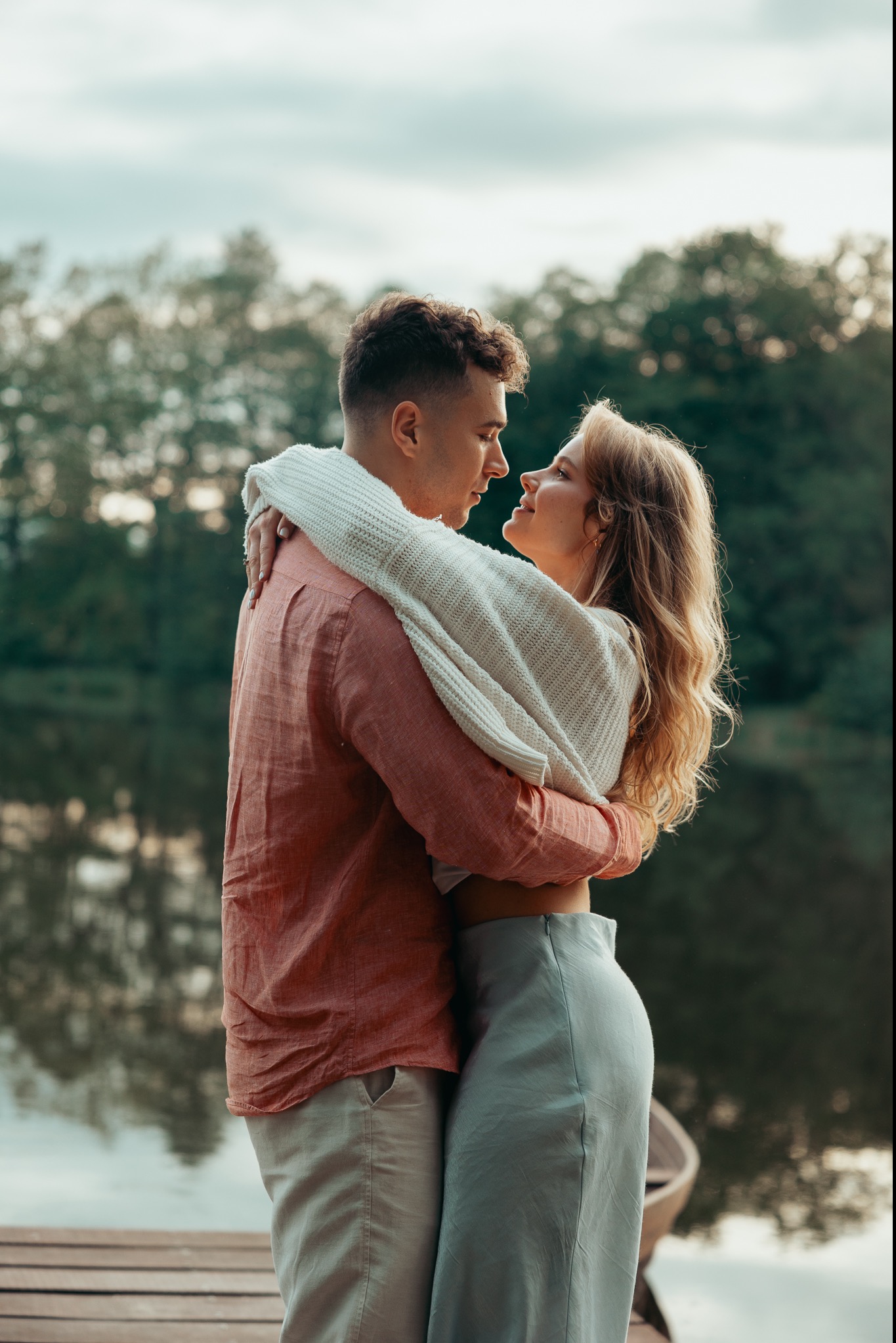 Para zakochanych obejmuje się na drewnianym pomoście nad jeziorem, ona w jasnej spódnicy i swetrze, on w koralowej koszuli.