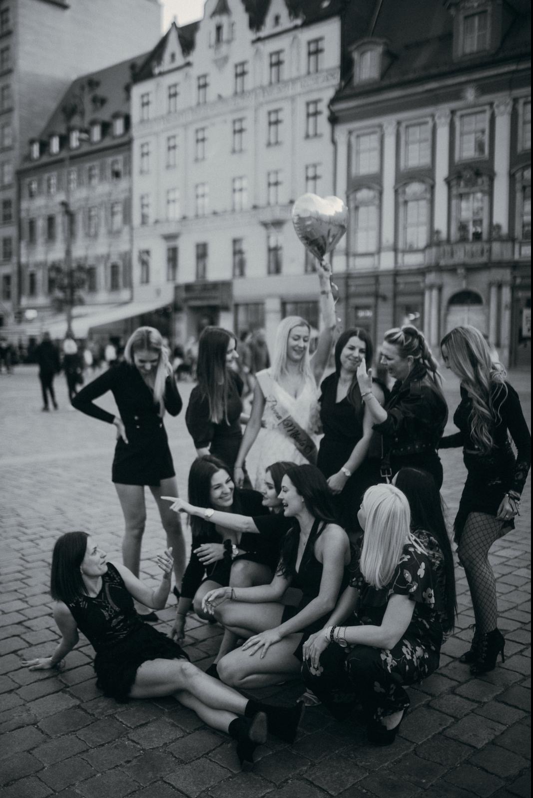 Czarno-białe zdjęcie grupy kobiet świętujących wieczór panieński na tle architektury miejskiej, panna młoda trzyma balon w kształcie serca.