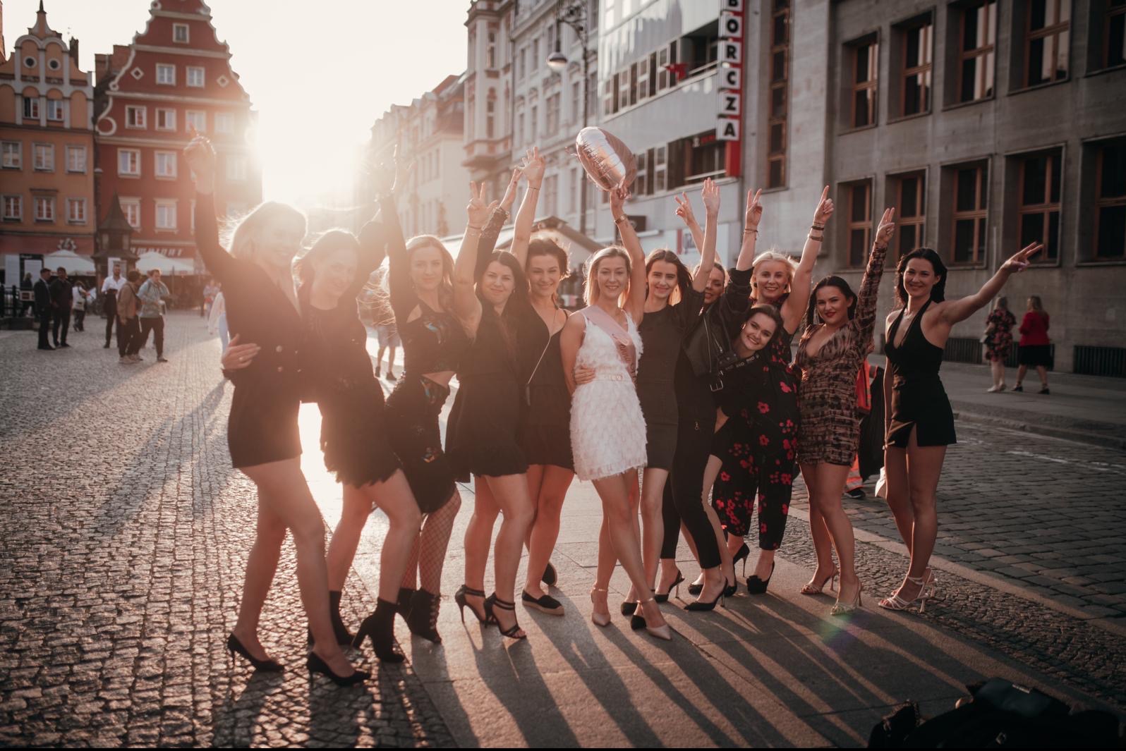 Grupa kobiet świętuje na brukowanej ulicy miasta, w tle budynki w stylu kamienic, jedna z kobiet ma szarfę i trzyma balon
