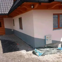 Ocieplona elewacja domu jednorodzinnego z pomalowaną dolną częścią na szaro, okna zabezpieczone folią, teren wokół domu wysypany kamieniami.