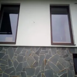 Fragment elewacji budynku z dwoma oknami o brązowych ramach i wykończeniem z nieregularnych kamiennych płyt w odcieniach szarości i brązu.