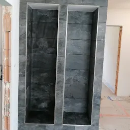 Dwa wnęki w ścianie wyłożone szarymi, poziomymi płytkami imitującymi kamień, obramowane srebrną listwą, w surowym stanie wykończenia.