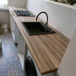 Kątowe ujęcie blatu kuchennego z drewnopodobnym wzorem, czarnym zlewem i kuchenką gazową, z pralką w zabudowie pod blatem, na tle wzorzystej podłogi i białych szafek w trakcie montażu.