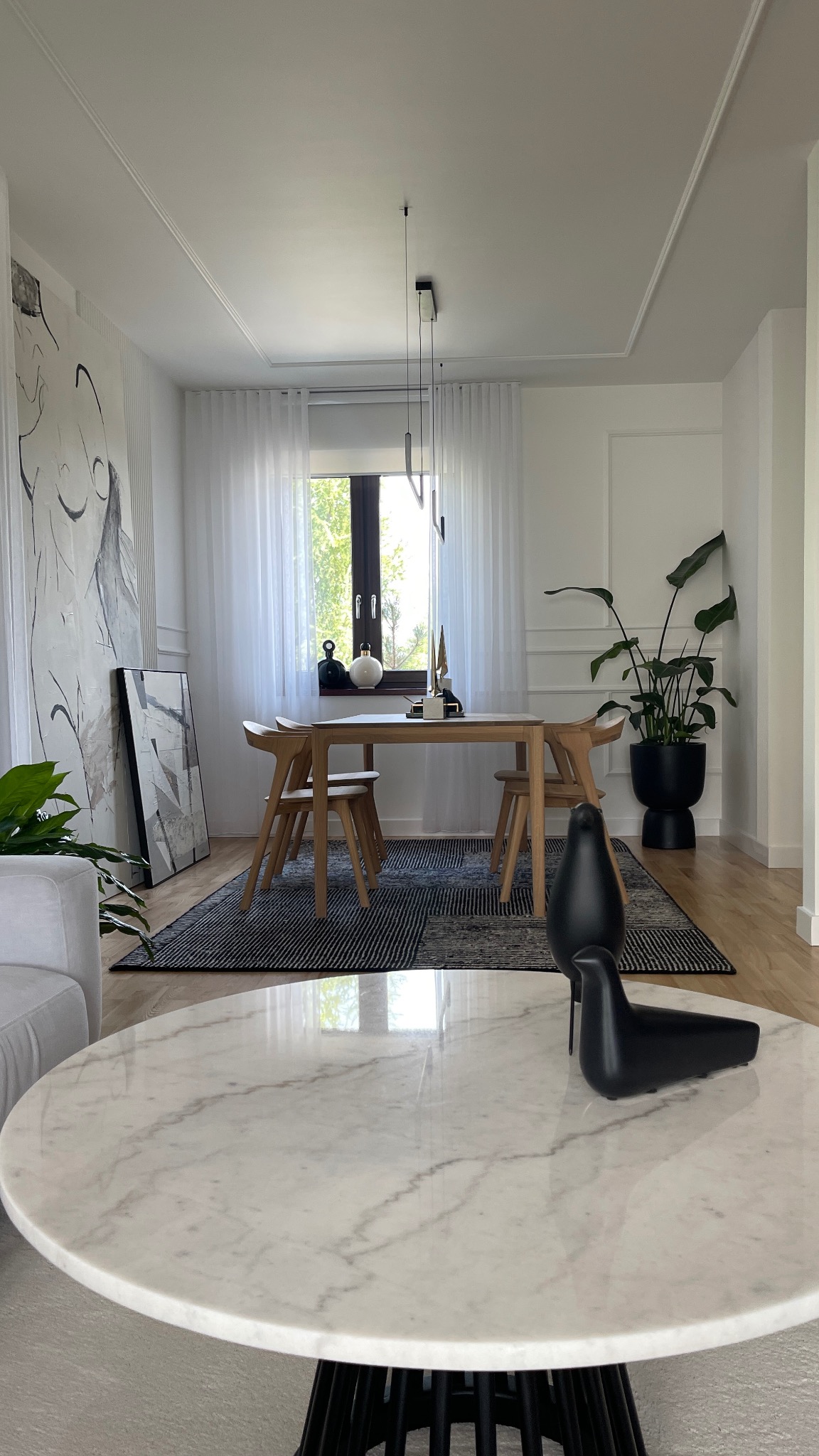 Jasne wnętrze jadalni z drewnianym stołem i krzesłami, czarnym dywanem w geometryczne wzory. Na pierwszym planie marmurowy stolik kawowy z dekoracyjnymi figurkami ptaków.