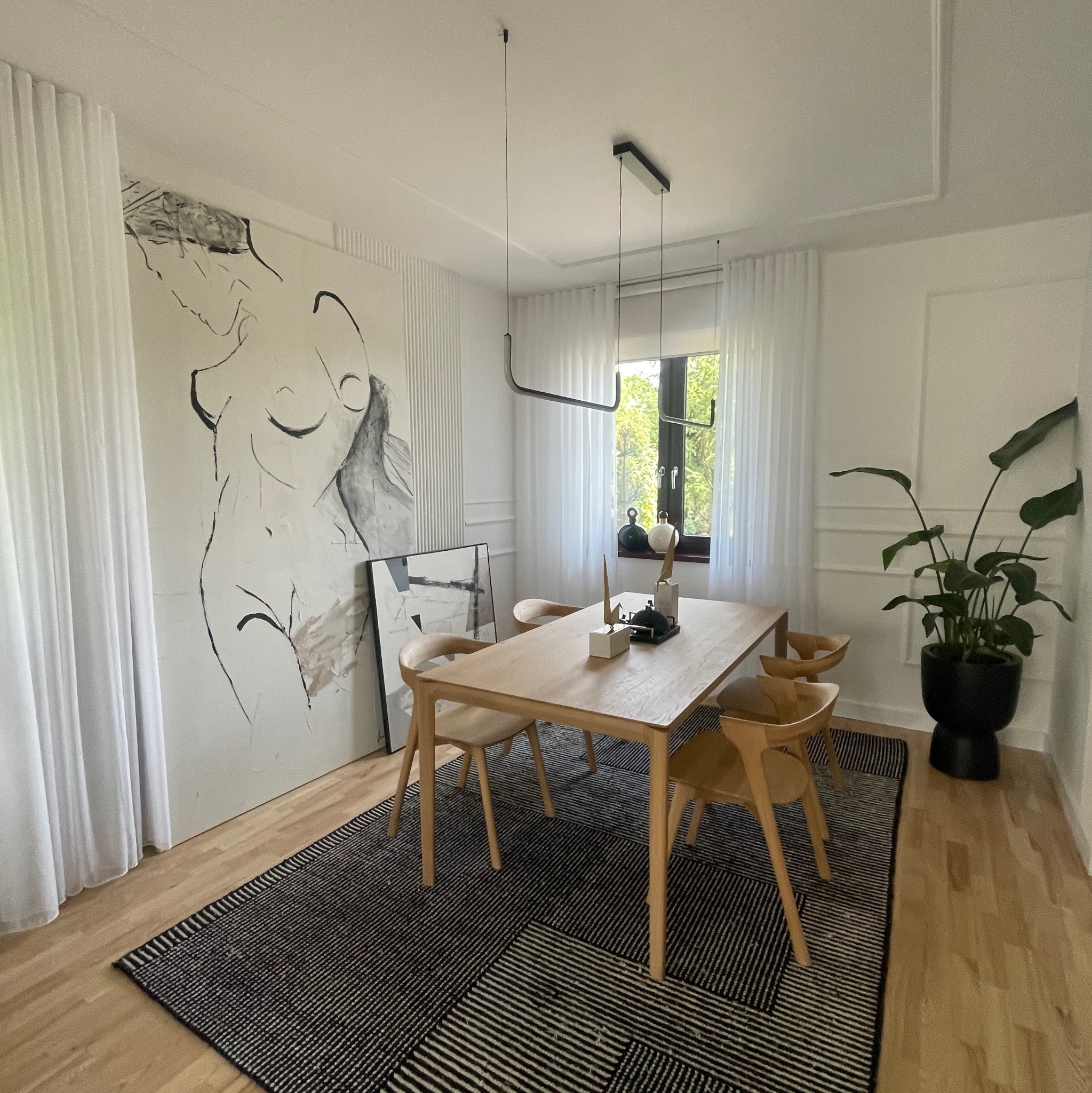 Jasna jadalnia z drewnianym stołem, krzesłami i czarno-białym dywanem. Na ścianie duży obraz. Minimalistyczne oświetlenie i roślina doniczkowa.