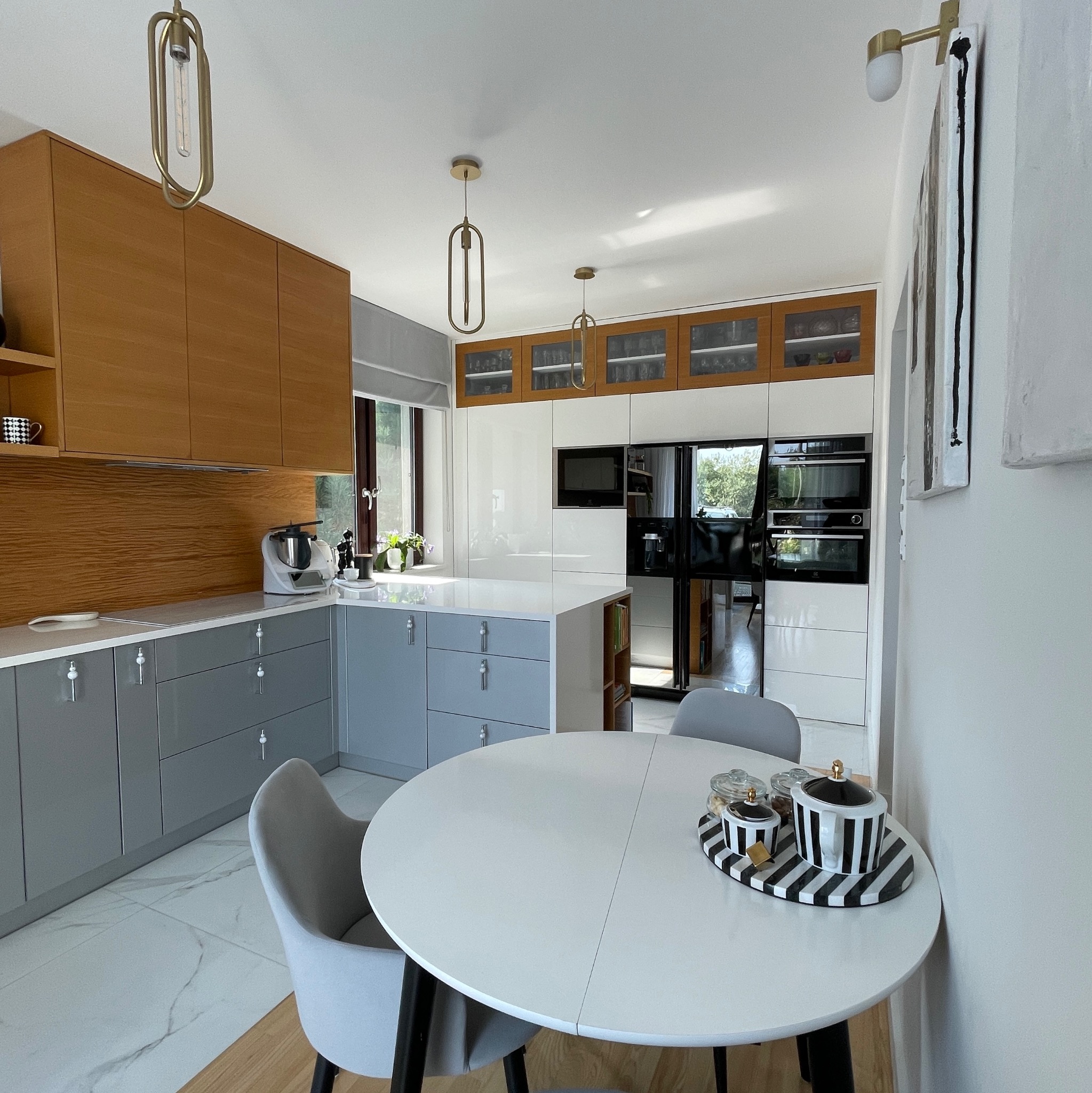 Nowoczesna kuchnia z drewnianymi i szarymi szafkami, białym blatem i okrągłym stołem. Widoczne oświetlenie i dekoracje w czarno-białe paski. Jasne wnętrze.