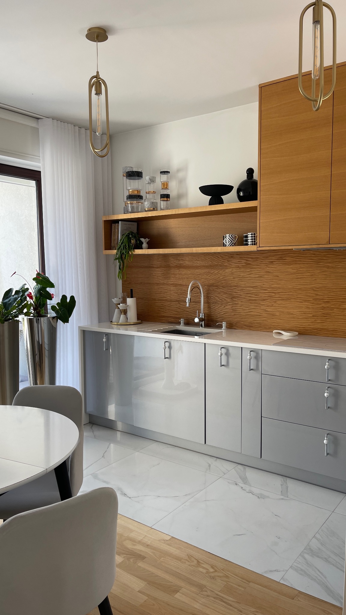 Nowoczesna kuchnia z szarymi szafkami, drewnianym panelem nad blatem i marmurową podłogą. Dwie lampy wiszące i rośliny doniczkowe dodają elegancji.
