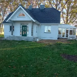 Parterowy dom z ciemną dachówką, przeszkloną werandą i zielonymi drzwiami wejściowymi, otoczony zielenią.