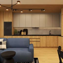 Magda Ciurkot Architektura Wnętrz - Salon z aneksem kuchennym w ciepłych barwach: drewniane szafki, szara ściana, niebieska sofa i czarne akcenty oświetleniowe. Nowoczesny design.