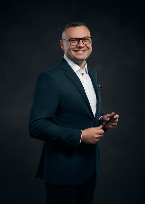 Uśmiechnięty mężczyzna w ciemnozielonym garniturze i okularach, trzymający czarny długopis na ciemnoszarym tle.