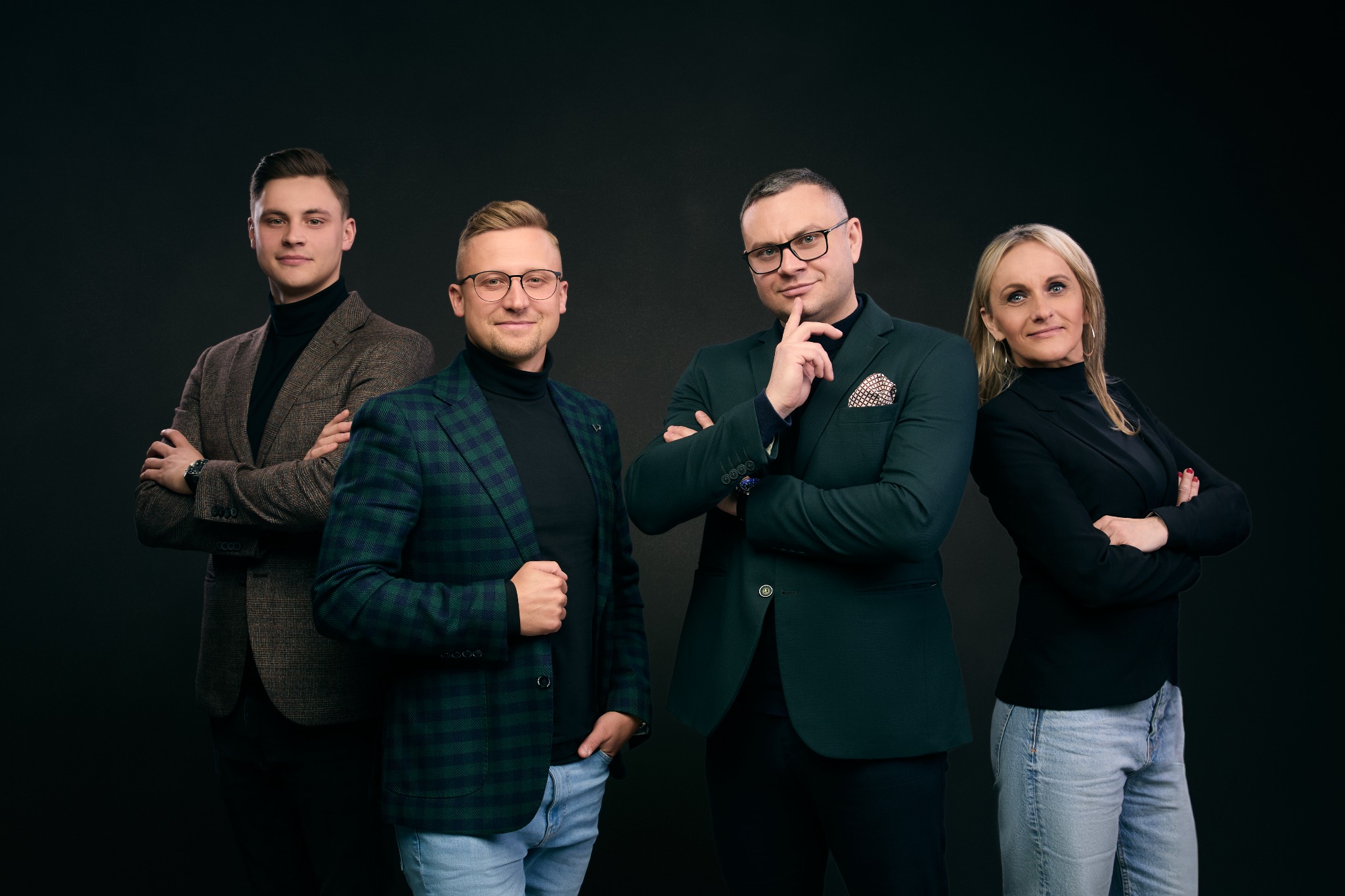 Zespół czterech osób w eleganckich marynarkach pozuje na ciemnym tle: mężczyzna w brązowym tweedzie, mężczyzna w zielono-niebieskiej kracie, mężczyzna z chusteczką w kieszonce i kobieta w czarnej...