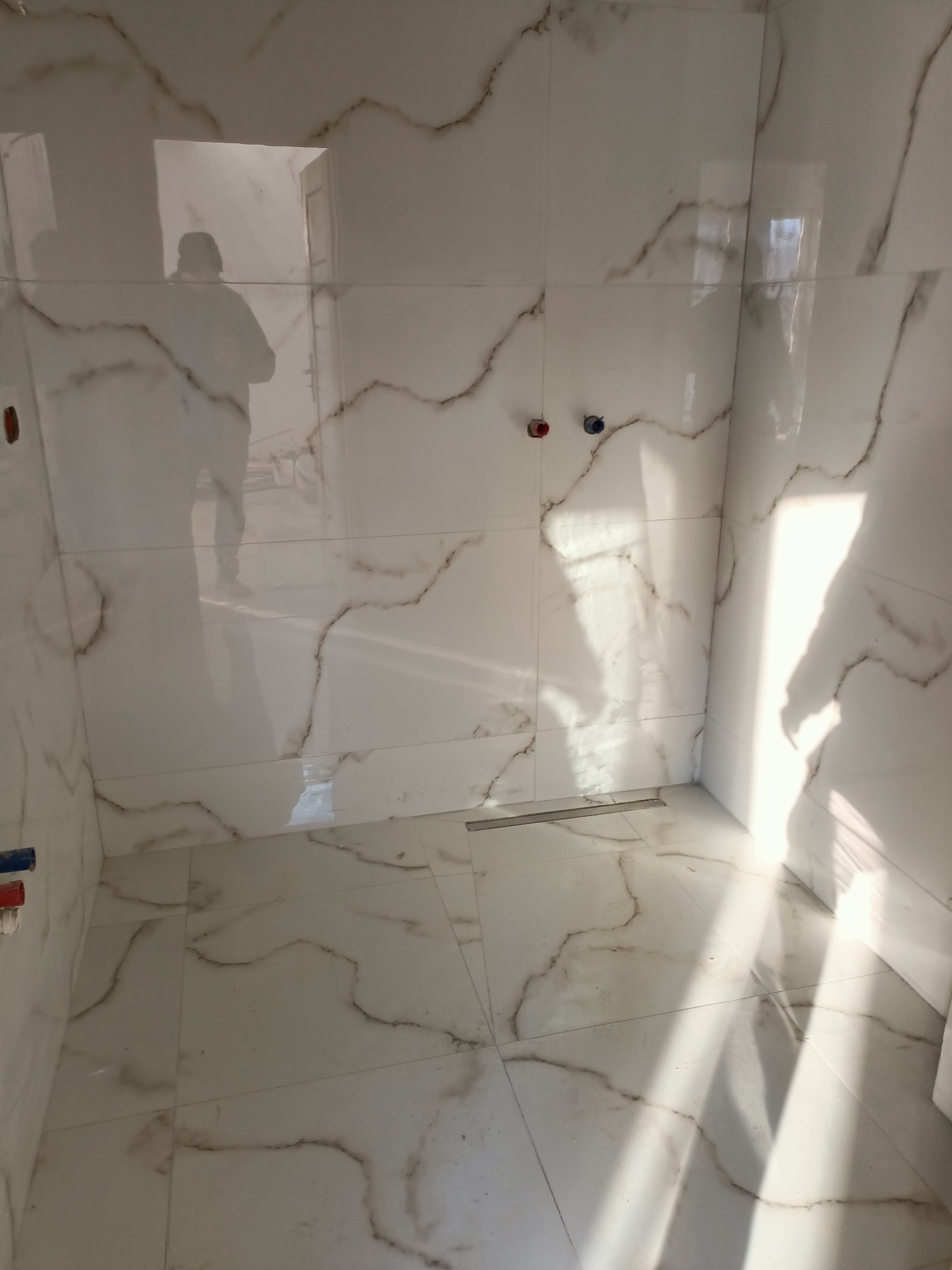 Nowoczesna łazienka w trakcie wykańczania, białe płytki z marmurkowym wzorem, widoczny odpływ liniowy i instalacja wodna, odbicie sylwetki osoby w płytkach.