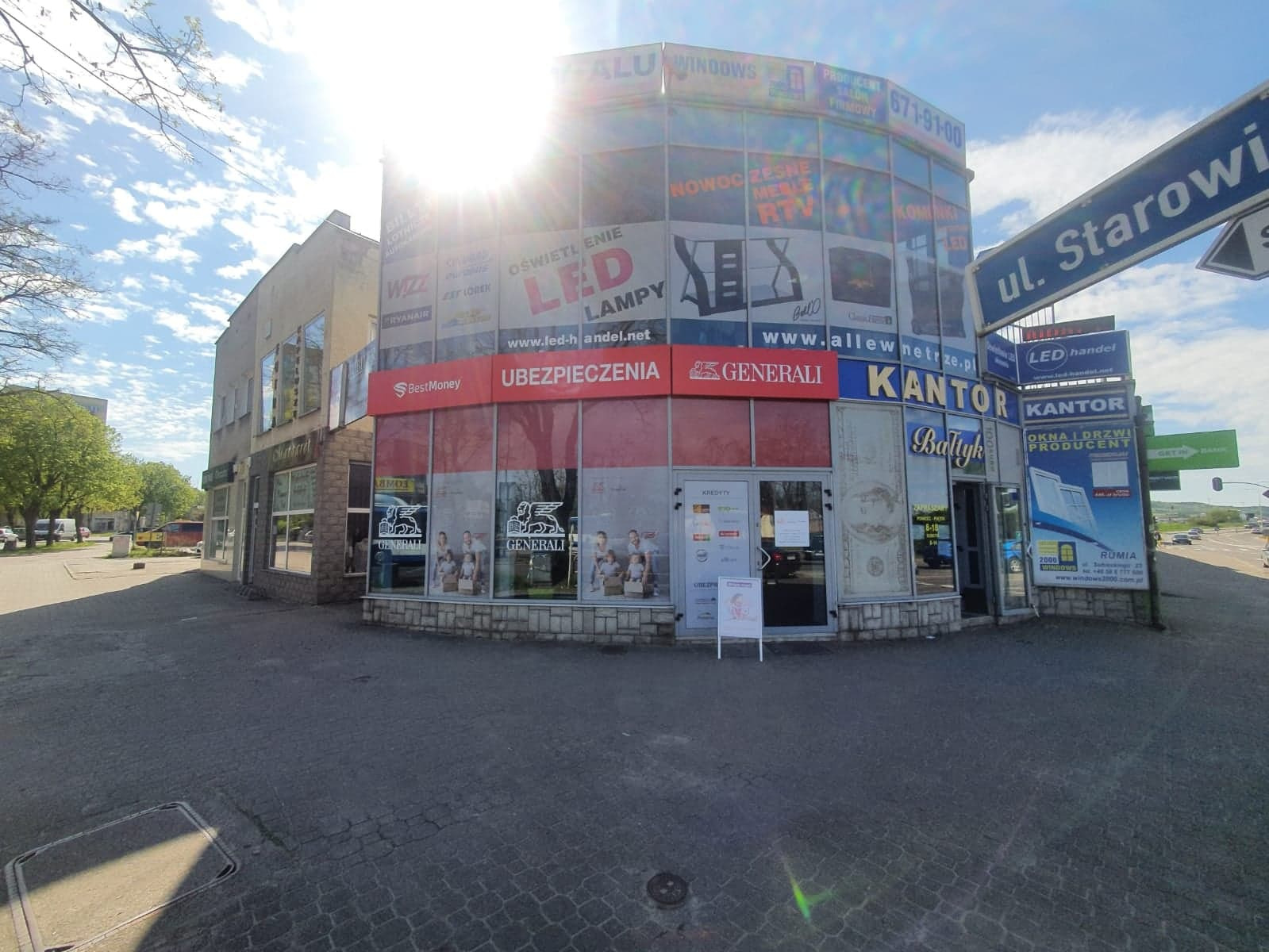 Budynek z reklamami, w tym szyldy BestMoney i Generali Ubezpieczenia nad wejściem, widok z zewnątrz w słoneczny dzień.