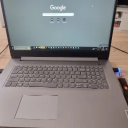 Szary laptop Lenovo z otwartą przeglądarką Google Chrome na drewnianym biurku, widoczny pasek zadań z ikonami aplikacji i podłączony pendrive z brelokiem.